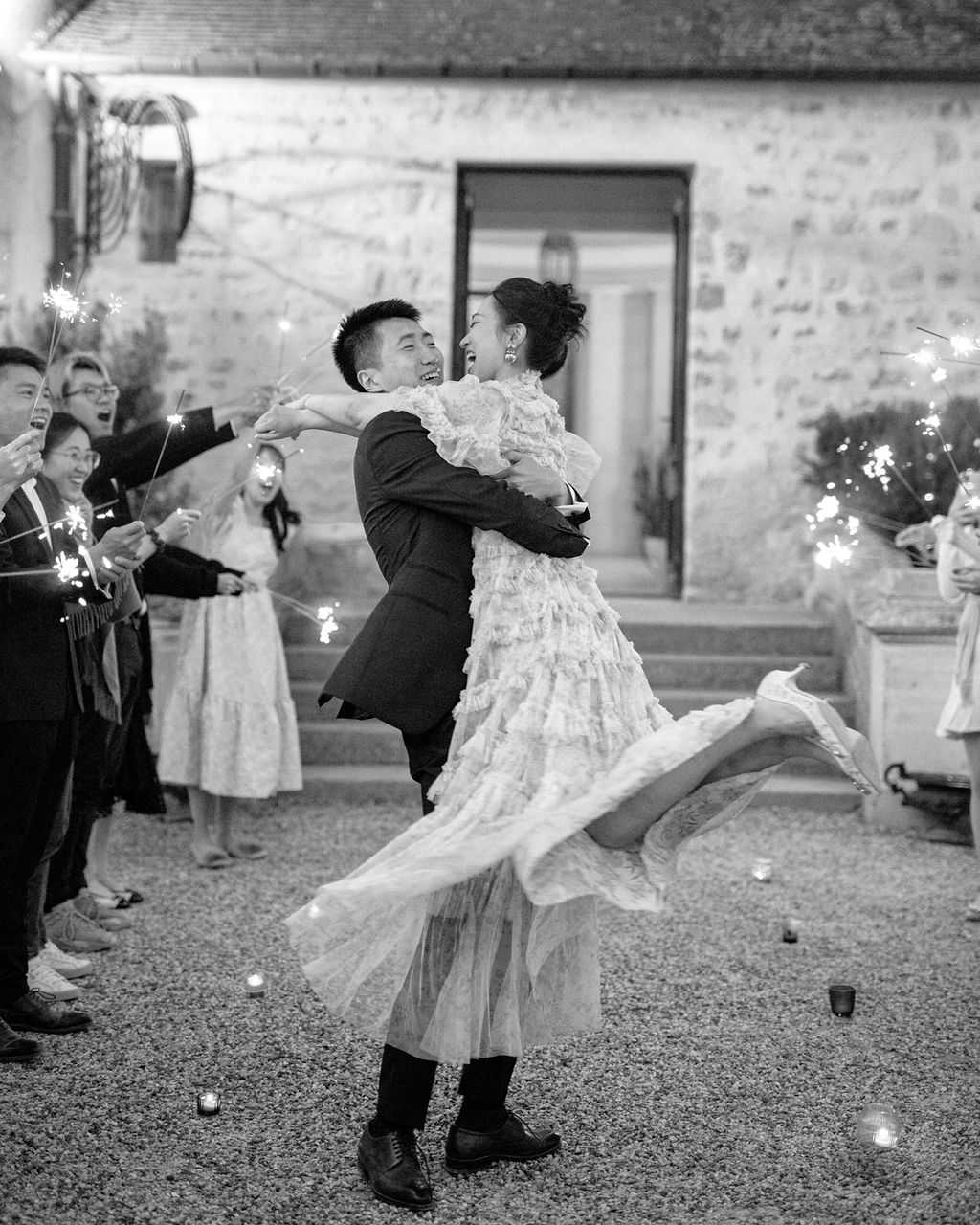 A black-and-white image of a sparkler send-off moment at an outdoor evening reception, set in a gravel courtyard in front of a stone building with steps and a doorway visible in the background. The groom, wearing a dark suit, is lifting and spinning the bride, who wears a tiered ruffled lace dress with a flowing skirt and her hair in an updo; both are laughing openly. Approximately eight to ten guests line both sides of the couple, holding lit sparklers that produce bright, high-contrast bursts of light against the dark background, while small votive candles are scattered on the gravel ground. The composition is a mid-distance portrait shot capturing the movement and energy of the moment with strong tonal contrast between the bright sparkler light and the dark surroundings.