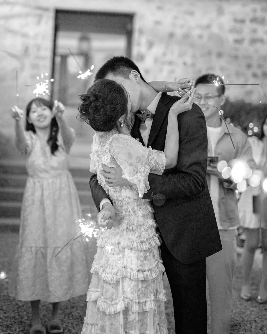 This black-and-white image captures a couple kissing during a sparkler send-off, shot in portrait format with the couple centered in the foreground. The bride wears a ruffled, tiered dress with floral detailing and long sleeves, her dark hair styled in an updo, and she holds a lit sparkler in one hand. The groom is dressed in a dark suit with a bow tie and has his arm wrapped around the bride. Several guests visible in the soft-focus background are holding lit sparklers and smiling, including a woman in a midi dress to the left. The setting appears to be an outdoor courtyard in front of a stone building, photographed at night with the sparklers providing bright, glowing light sources against the dark tones of the image.