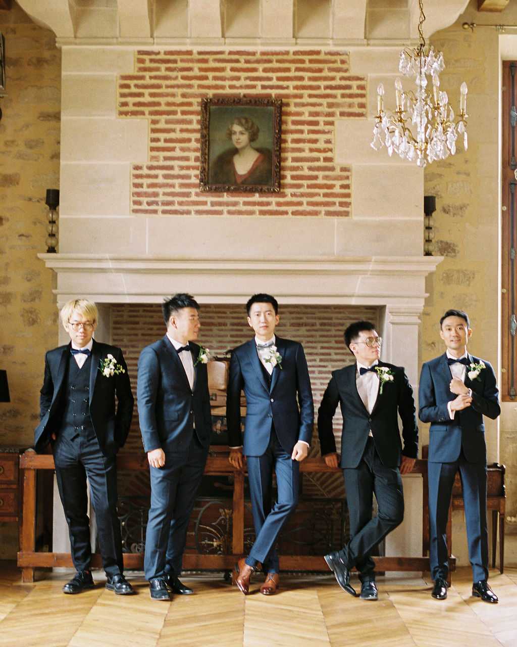 A groom and four groomsmen pose casually inside a chateau, standing in front of a large stone fireplace with exposed brick interior and a gilt-framed portrait hanging above the mantel. The room features a crystal chandelier, parquet flooring, and stone walls, conveying a classic French chateau interior. The groom, positioned at center, wears a navy three-piece suit with a patterned bow tie and white boutonniere, while the groomsmen wear navy and black tuxedo suits with black bow ties and matching white boutonnieres with greenery. This is a wide group portrait shot with the five men arranged in a relaxed, candid-style stance.