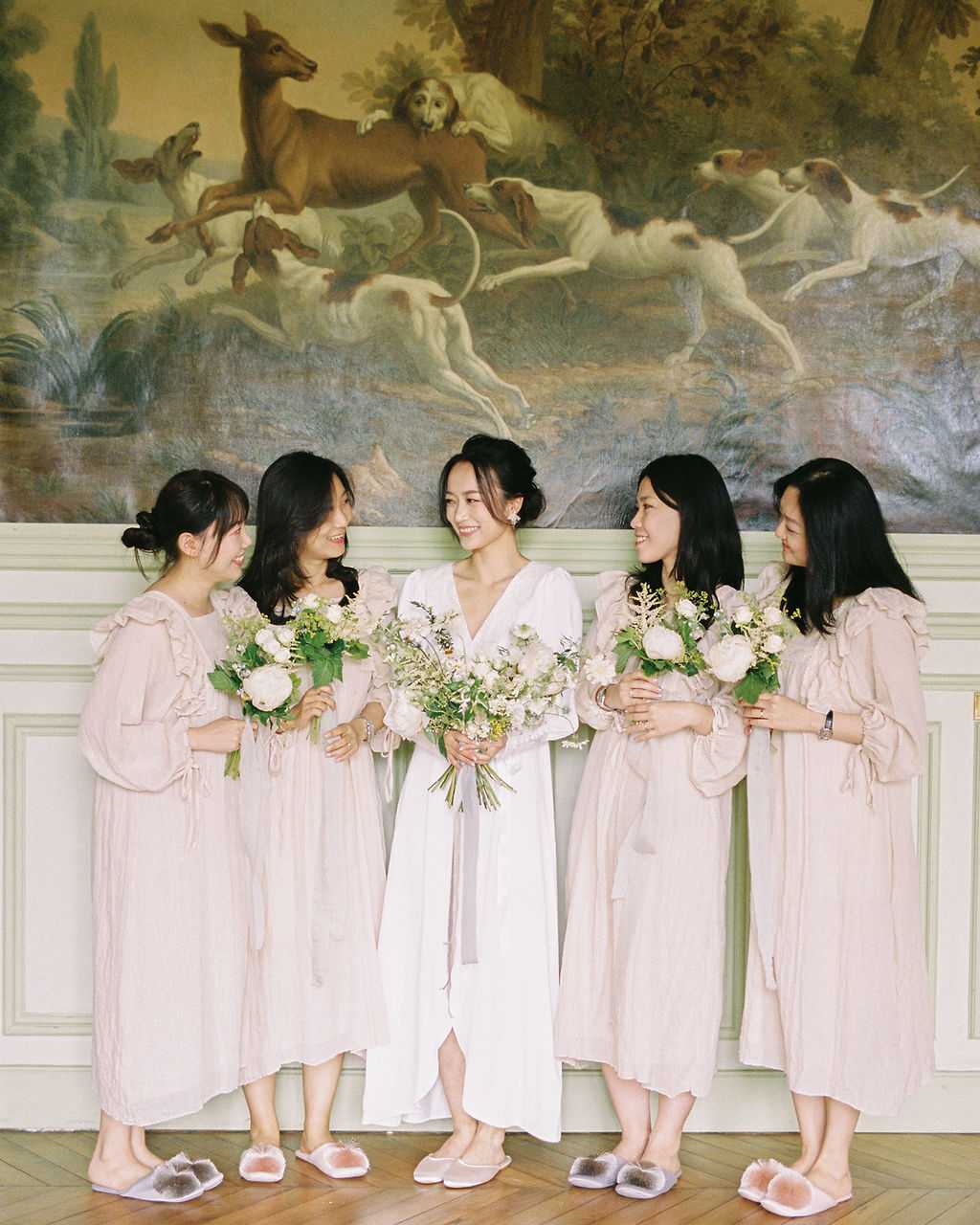 A getting-ready portrait of a bride standing with four bridesmaids inside what appears to be a French chateau, posed in front of a large classical oil painting depicting a hunting scene with dogs and deer. The bride wears a white long-sleeve V-neck wrap-style dress with an upswept hairstyle and pearl drop earrings, holding a loose garden-style bouquet of white peonies, ranunculus, and mixed greenery. The four bridesmaids wear matching blush pink long-sleeve midi dresses with ruffle neckline details, each holding a smaller version of the same bouquet, and all five women wear matching fluffy pompom flat slippers in pale lavender-gray. The group is laughing and looking at one another in a relaxed, candid moment, captured in a medium full-length portrait shot with film grain visible, giving the image a soft, warm tone.