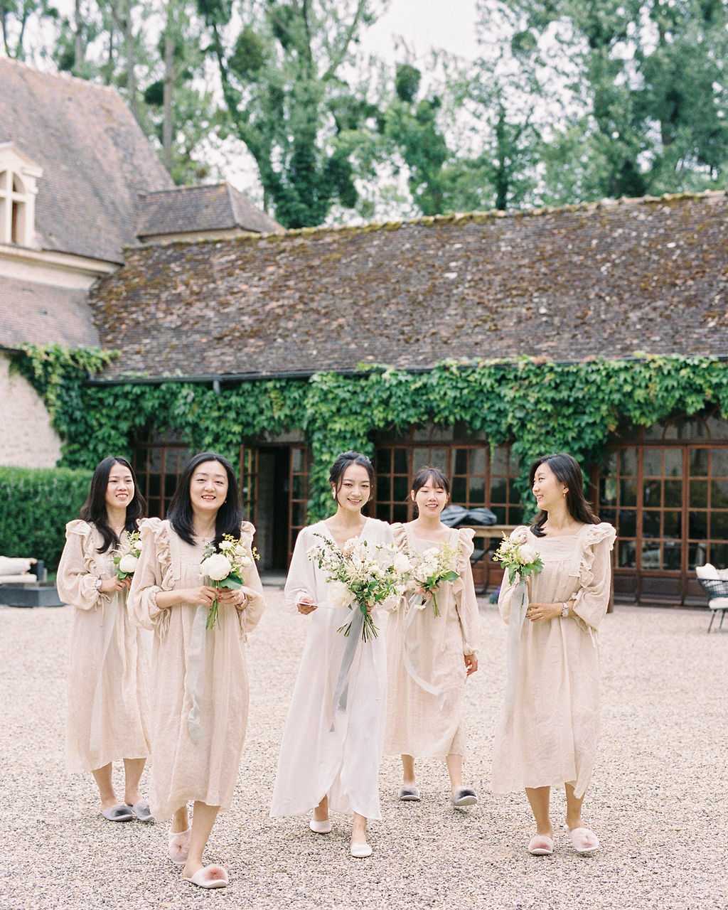 A bridal party of five women walks across a gravel courtyard at a French chateau or manor property, shot as a medium wide portrait. The bride is centered, wearing a white dress with a veil and white flat shoes, and carries a loose bouquet of white peonies, small yellow blooms, and greenery. The four bridesmaids wear matching midi-length linen dresses in a warm sand-beige tone with ruffled shoulder detailing and long sleeves, paired with casual fluffy slippers in grey and blush tones. Each bridesmaid holds a smaller bouquet of white flowers and greenery consistent with the bride's arrangement. The styling is understated and minimal, with a soft, natural palette. The background features a vine-covered stone outbuilding with wood-framed windows typical of a French estate courtyard setting.
