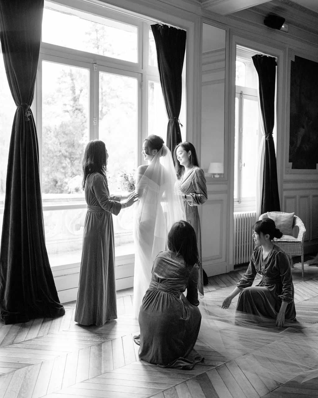 This black-and-white image captures a getting-ready moment inside a chateau or manor house, where a bride in a fitted white gown with a long veil and an updo is surrounded by four bridesmaids in floor-length velvet dresses. Two bridesmaids stand on either side of the bride near tall French windows with heavy draped curtains, one holding her hands, while two others kneel on the herringbone parquet floor arranging her veil. The room features classic boiserie wall paneling, a decorative radiator, and a white wicker chair, indicating a formal historic interior. The image is shot in a wide portrait format with strong backlighting from the windows creating bright highlights and deep shadow contrast, and the bridesmaids' velvet dresses render as a rich mid-to-dark gray tone throughout.