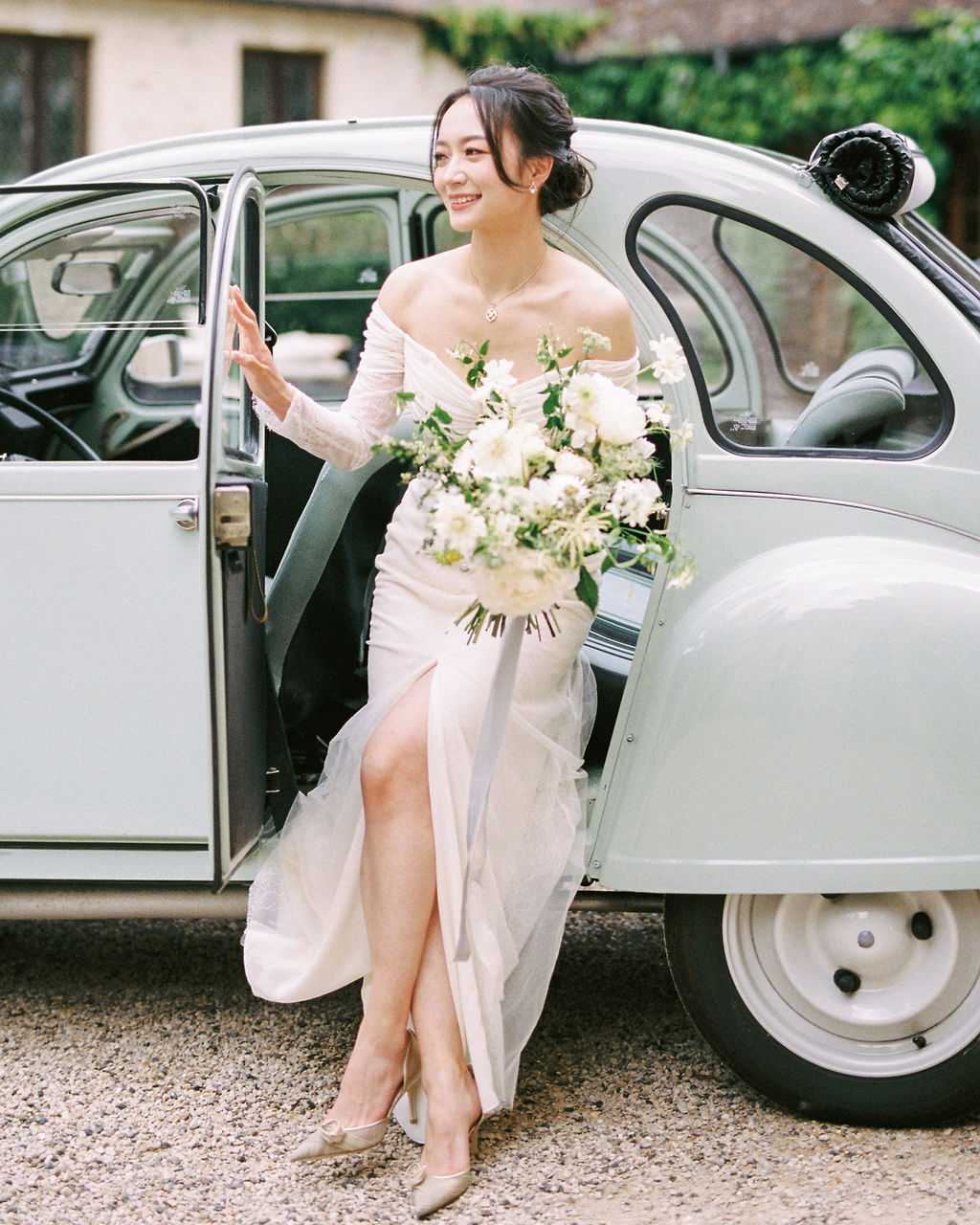 A bride steps out of a vintage mint-green Citroën 2CV on a gravel courtyard, holding the open car door with one hand and a large bouquet in the other. She wears an off-the-shoulder long-sleeve ivory gown with a front slit and a draped bodice, paired with champagne-colored heeled pumps and a delicate pendant necklace, with her dark hair styled in a loose updo. The bouquet is a lush, garden-style arrangement of white dahlias, white cosmos, and greenery. The shot is a medium full-length portrait taken outdoors, with a stone building and ivy visible in the background.
