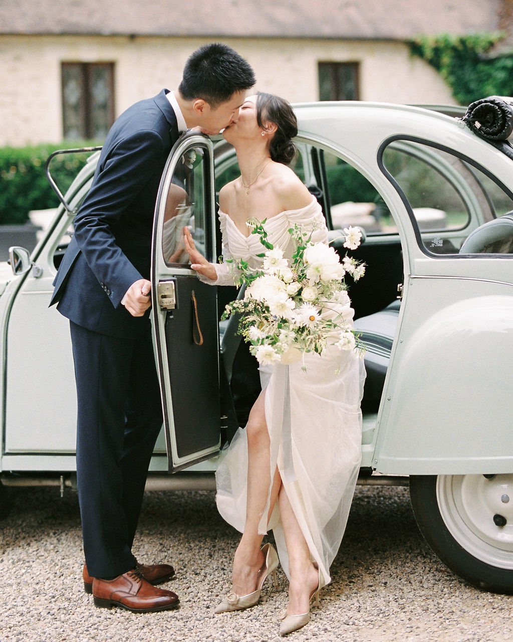 A couple portrait shot outdoors on a gravel courtyard, with the bride seated in the open door of a vintage pale sage green Citroën 2CV as the groom leans in to kiss her. The groom wears a navy suit with brown leather oxford shoes, while the bride wears an off-the-shoulder ivory draped gown with a front slit and champagne heeled mules. She holds a large loosely arranged bouquet of white ranunculus, white cosmos, white garden roses, and trailing greenery. The background features a stone building facade with ivy, consistent with a French chateau or manor property. The image has a film photography quality with soft, warm tones and a mid-range portrait composition.