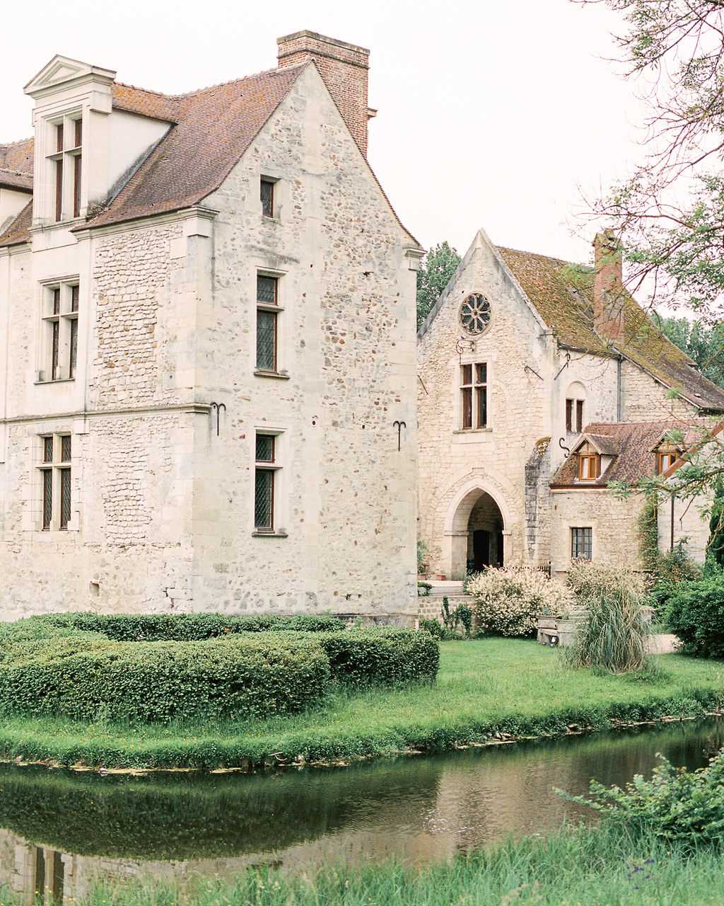 Wide exterior shot of a historic French stone chateau featuring medieval architecture, including a Gothic arched entrance doorway, a rose window above the entry, terracotta-tiled rooflines, and multi-pane leaded windows. The building complex includes several connected structures of varying heights, all constructed from pale limestone. A still moat or pond reflects the facade in the foreground, bordered by neatly trimmed low hedging and green lawn. No people are visible in the frame. The image has a soft, slightly muted film quality. Potential venue feature image.