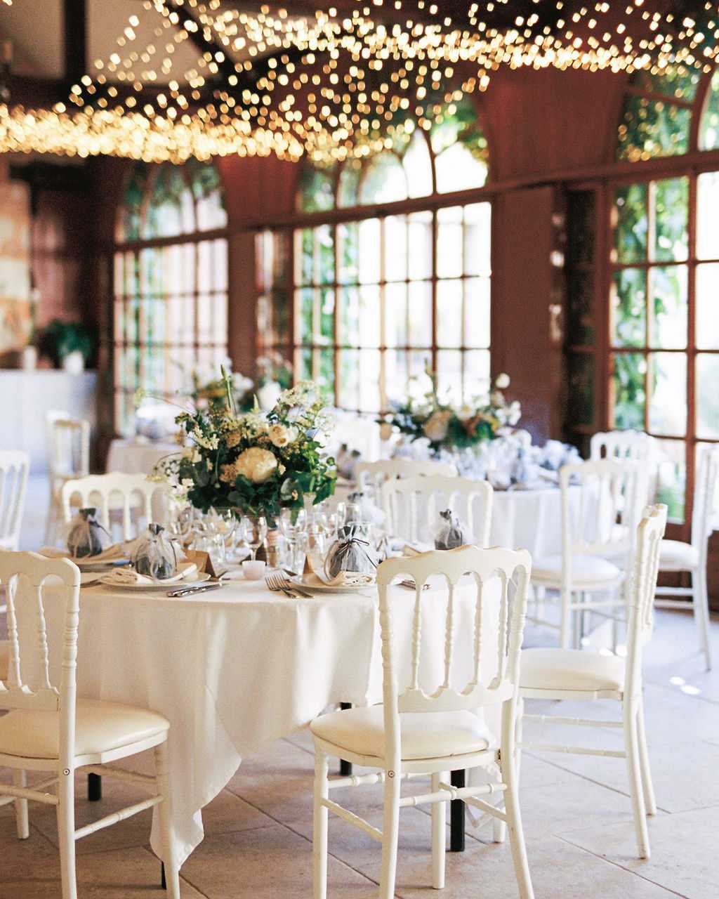 A wedding reception setup shot inside a glass-windowed orangery or conservatory-style venue, with no guests present. Multiple round tables are dressed in white floor-length linens and surrounded by white Napoleon-style chairs with cream cushioned seats. The centerpieces feature low, lush arrangements of ivory peonies, white blooms, and green foliage in clear glass vessels. Place settings include glassware, silverware, kraft paper or neutral-toned napkin folds, and small organza favor bags tied with ribbon. Overhead, a dense canopy of warm fairy lights covers the entire ceiling, creating a bokeh-heavy golden glow above the room. The venue's large arched multi-pane windows with dark wood frames allow natural daylight in and reveal greenery outside. The overall decor palette is white, ivory, and green with a classic French style. Wide interior shot.