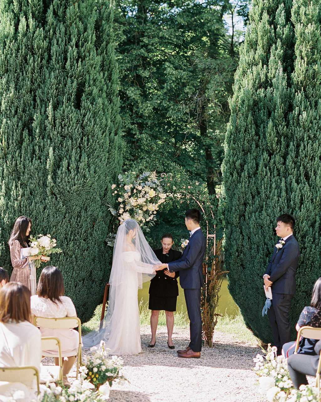 An outdoor wedding ceremony taking place on a gravel path flanked by tall, closely trimmed cypress hedges forming a natural corridor. The bride wears a long-sleeved ivory off-shoulder gown with a cathedral-length veil, and the groom is dressed in a navy suit; they are holding hands in front of an officiant in a black dress who stands before a circular metal arch adorned with white and soft yellow florals with eucalyptus greenery. A bridesmaid in a dusty mauve dress stands to the left holding a white floral bouquet, while a groomsman in a navy suit stands to the right. Seated guests, approximately 10-15 visible, are arranged on gold chairs on either side of the aisle, with loose clusters of white and yellow florals placed at ground level along the aisle edges. The composition is a medium-wide shot taken from behind the seated guests, capturing the full ceremony scene in a classic, garden-formal style.