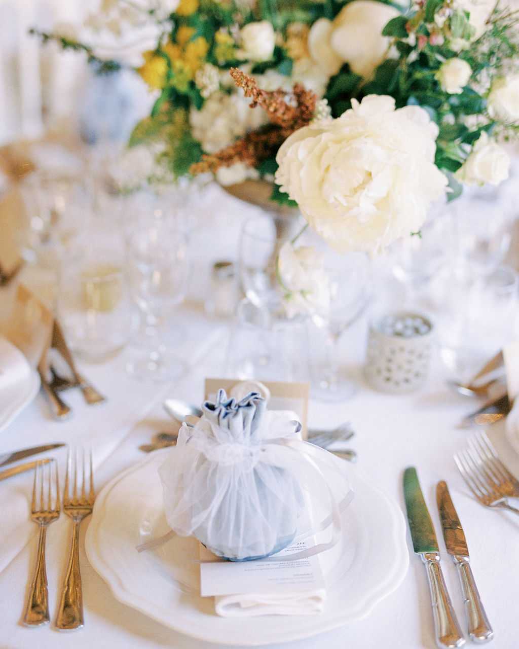 Close-up detail shot of a wedding reception place setting on a white linen tablecloth. The place setting features white china plates layered on a charger, silver cutlery with decorative handles, and a small pale blue velvet drawstring pouch serving as a guest favor, placed on top of a printed menu card. In the background, a lush centerpiece arrangement includes ivory peonies, yellow blooms, dried rust-colored foliage, and greenery, along with crystal glassware and a small patterned votive candle holder. The overall decor palette is white, soft blue, and yellow with gold accents, suggesting a classic, airy reception style.