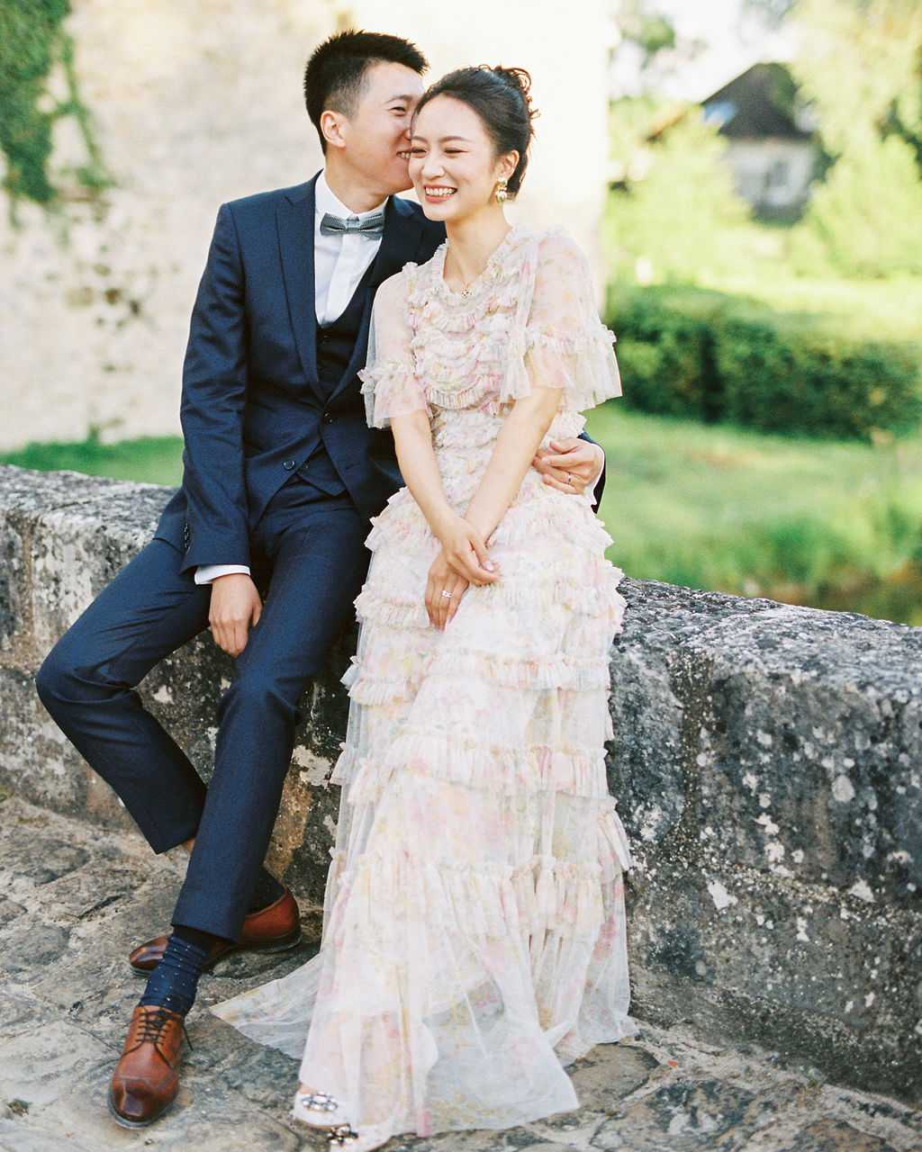 A couple portrait shot outdoors, with the pair seated on a low stone wall. The groom wears a navy three-piece suit with a grey bow tie and brown leather oxford shoes, and leans in to kiss the bride on the cheek. The bride is laughing and wears a floor-length tiered ruffle gown in blush with a soft floral print, flutter sleeves, and embellished flat shoes; her dark hair is styled in an updo. The setting appears to be the grounds of a historic stone building, with greenery and a structure visible in the soft-focus background. The image has a film photography quality with warm, slightly faded tones consistent with analog shooting.