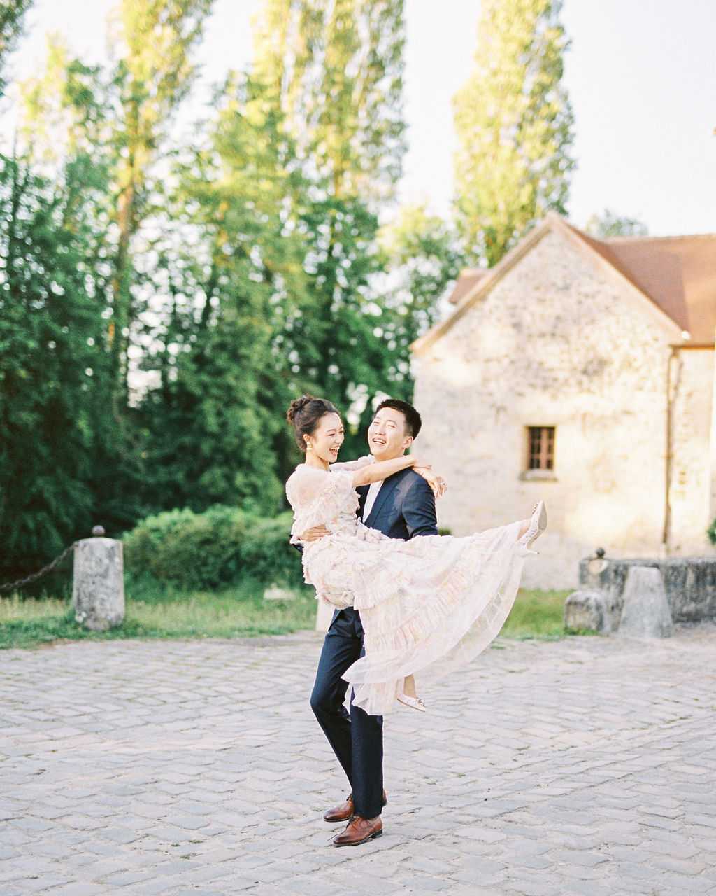 A couple portrait shot outdoors on a cobblestone courtyard, with the groom lifting the bride as both laugh openly. The groom wears a navy suit with brown leather oxford shoes, while the bride wears a blush pink tiered ruffle dress with sheer long sleeves and her hair in an updo. The setting features a French stone building with an orange-tiled roof in the background, shot during warm golden hour light. The image is a medium full-body portrait with a softly blurred background.