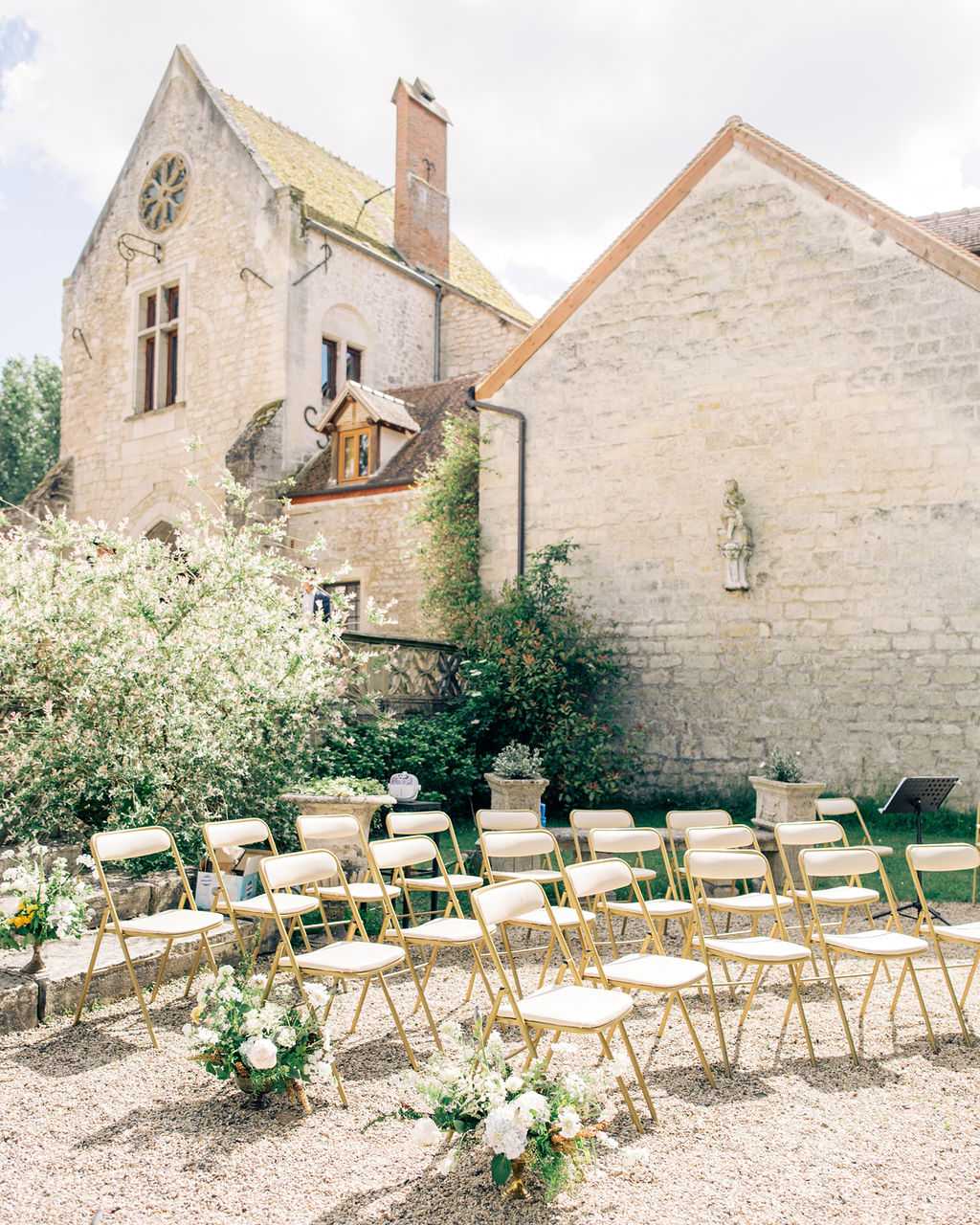 An outdoor ceremony setup in the courtyard of a historic French stone building, likely a chateau or abbey, featuring a rose window and a carved stone wall niche with a religious statue. Approximately 24 gold-framed folding chairs with cream upholstered seats are arranged in rows on a gravel surface, divided by a central aisle. White and cream floral clusters with greenery mark the ends of the aisle rows at ground level. A music stand is visible to the right, and potted topiaries flank the ceremony area. No guests or wedding party are present — the space is set up and awaiting the ceremony. Wide-angle shot capturing both the seating arrangement and the full facade of the venue. Potential venue feature image.