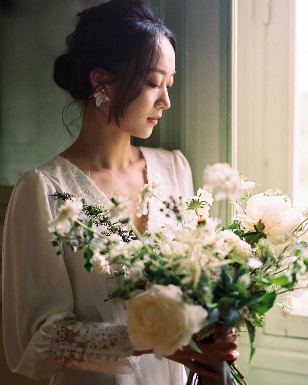 A bride stands indoors near a window during a getting-ready moment, looking downward at her bouquet in soft natural side light. She wears a white long-sleeve dress with a lace collar and lace cuff detail, her dark hair styled in a loose updo with face-framing tendrils, and she accessorizes with delicate floral drop earrings. The bouquet is a large, loosely gathered arrangement of cream peonies, white cosmos, scabiosa, astrantia, and trailing greenery with dark thistle accents, giving it an organic, garden-gathered aesthetic. The portrait is a close-up shot from roughly chest height, with a shallow depth of field that keeps the bride's face sharp against a softly blurred muted green background.