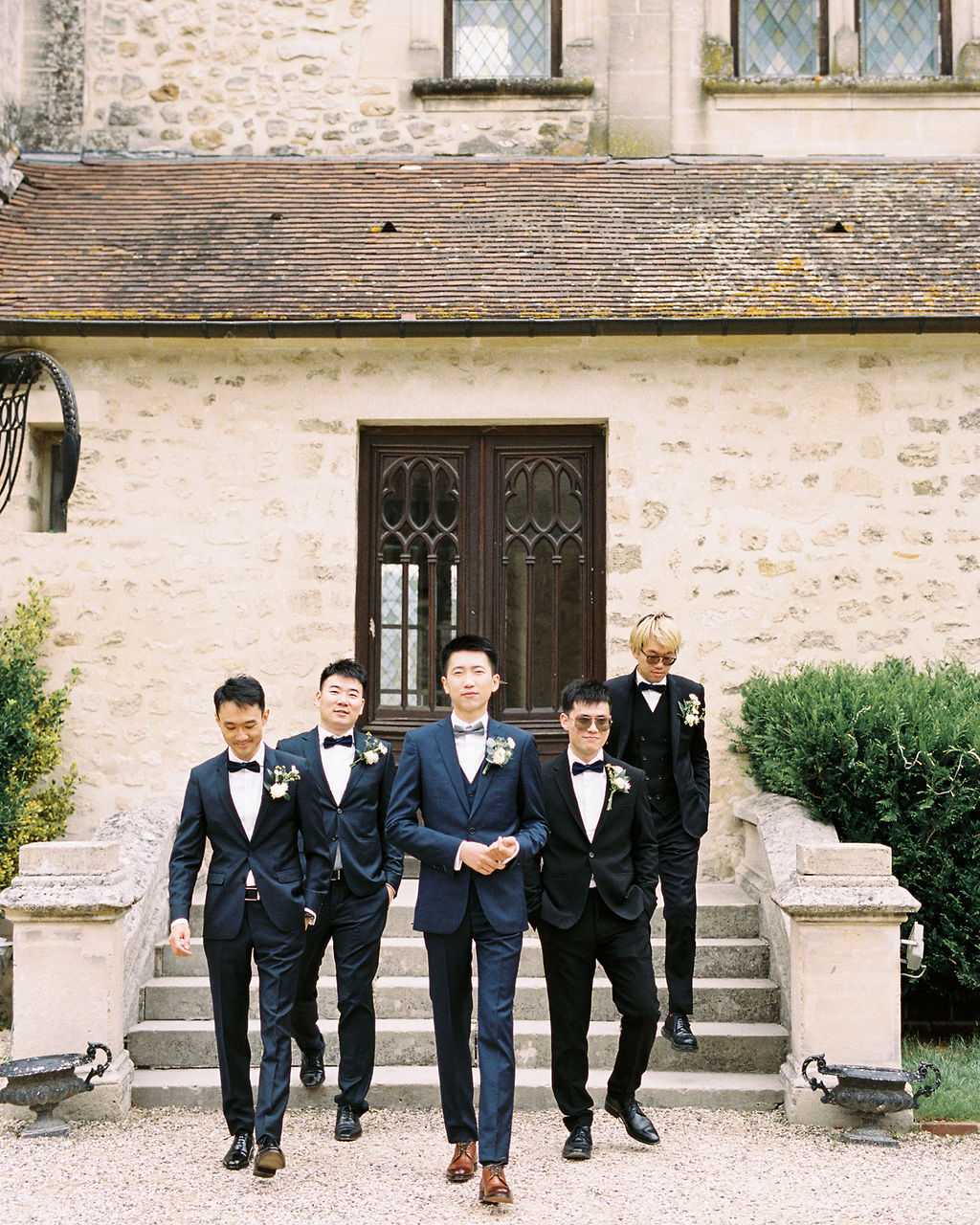 A groom and four groomsmen walk down the stone steps of a French chateau toward the camera in a wide portrait shot. The groom stands at the center front, dressed in a navy blue three-piece suit with a light grey bow tie and brown leather Oxford shoes, while the four groomsmen wear black suits with black bow ties and black dress shoes. All five men wear small white floral boutonnieres. Two of the groomsmen wear sunglasses, and one has bleached blonde hair and wears a black waistcoat. The group is positioned in front of the chateau's limestone facade, which features a Gothic-style arched wooden door and leaded windows, with a gravel courtyard in the foreground. The overall styling is classic and formal. Potential venue feature image.