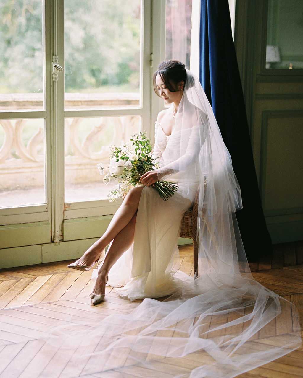 A bride portrait taken indoors, with the bride seated on a wicker chair beside tall French doors that open onto a stone balustrade. She wears an ivory long-sleeve fitted gown with a thigh-high slit and a cathedral-length tulle veil that pools extensively across the herringbone parquet floor. Her hair is styled in a low updo and she wears silver pointed-toe heels. She holds a loose, garden-style bouquet of white ranunculus, small white blooms, and trailing green foliage. The room features sage-green paneled walls and a deep navy curtain, and soft natural window light illuminates the scene from the left. The shot is a full-length portrait with a classic, refined editorial feel.