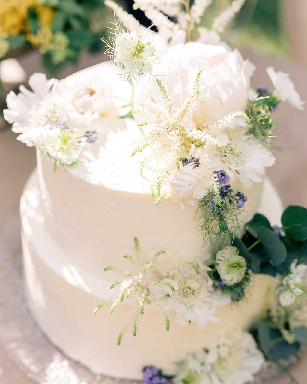 Close-up detail shot of a two-tier wedding cake with a smooth ivory buttercream finish, decorated with an abundant cascade of fresh flowers including blush peonies, white scabiosa, white daisies, nigella (love-in-a-mist) with its feathery green foliage, small purple forget-me-nots or veronica, eucalyptus leaves, and delicate green astilbe or astrantia sprigs. The floral decoration runs from the base of the top tier down across the lower tier, creating an asymmetrical garden-style arrangement. The overall color palette is white, blush, soft purple, and green, consistent with a romantic garden or boho wedding aesthetic. The image is shot from a slightly elevated angle in bright natural light with a softly blurred background.