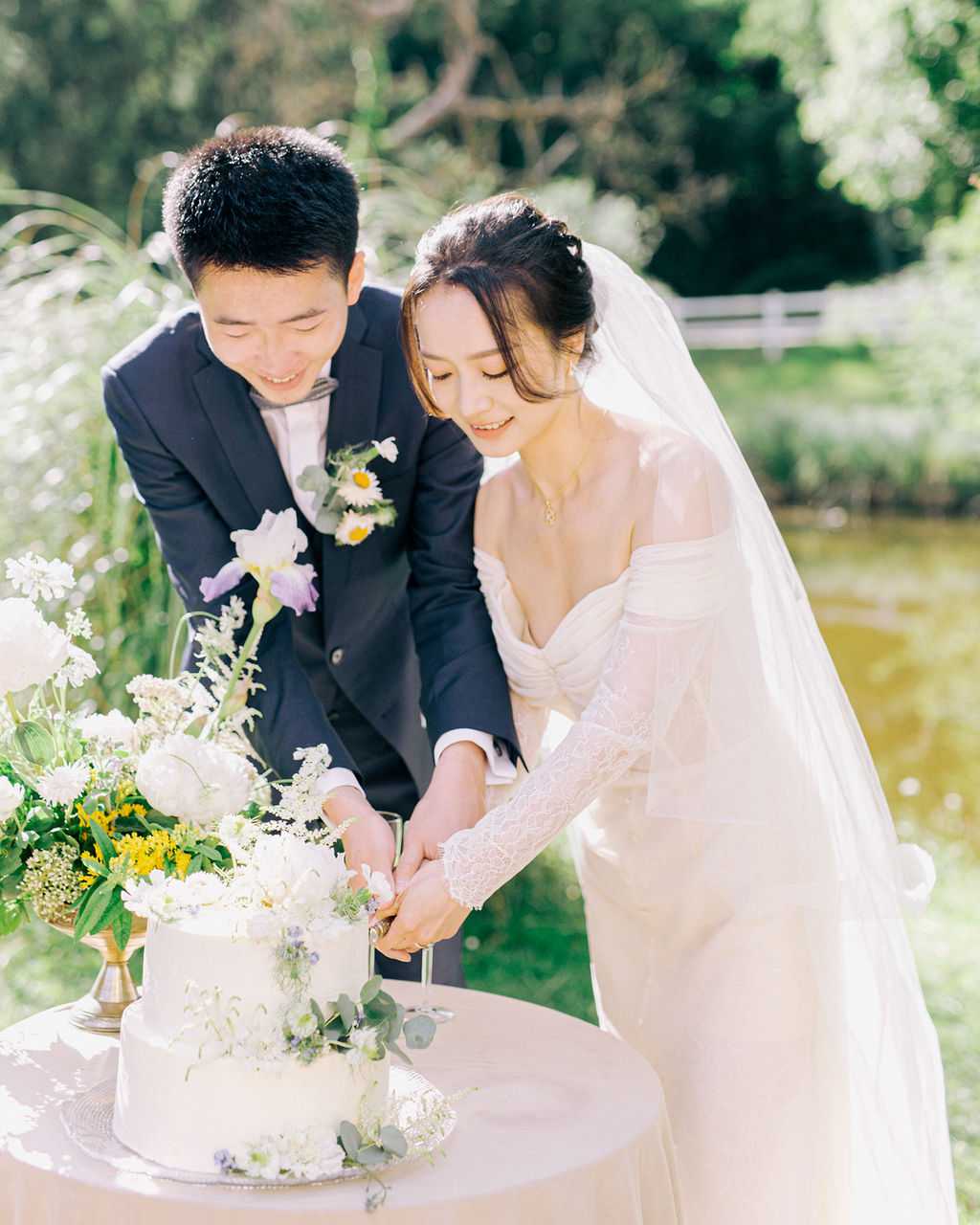 The bride and groom are cutting their wedding cake together at an outdoor garden setting, with a pond and white fence visible in the background. The groom wears a dark navy suit with a white daisy and greenery boutonniere, while the bride wears an off-the-shoulder ivory gown with long lace sleeves and a cathedral-length veil, her hair worn up with small floral pins. The two-tier white cake is decorated with white blooms, small purple flowers, and trailing eucalyptus, placed on a silver cake stand atop a blush-covered round table alongside a floral arrangement featuring lavender iris, white peonies, yellow accent flowers, and lush greenery in a gold compote vase. The composition is a medium portrait shot taken in bright natural light with a softly blurred background.