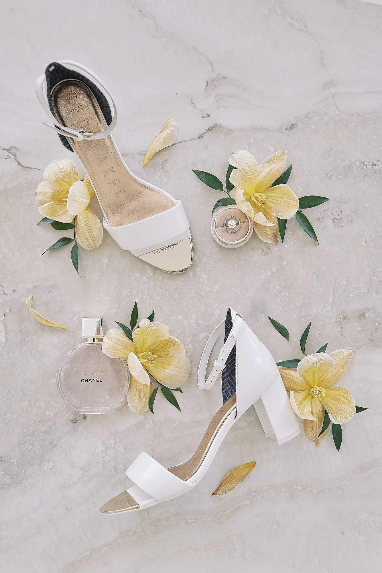 A flat lay detail shot of bridal accessories arranged on a white marble surface. The composition features a pair of white Ted Baker block-heel sandals with gold toe caps and ankle straps, a bottle of Chanel Chance perfume, and a diamond solitaire engagement ring in a nude ring box, all styled with scattered yellow tropical flowers and small green leaves. The overall palette is white, gold, and soft yellow, giving the arrangement a clean, modern aesthetic.