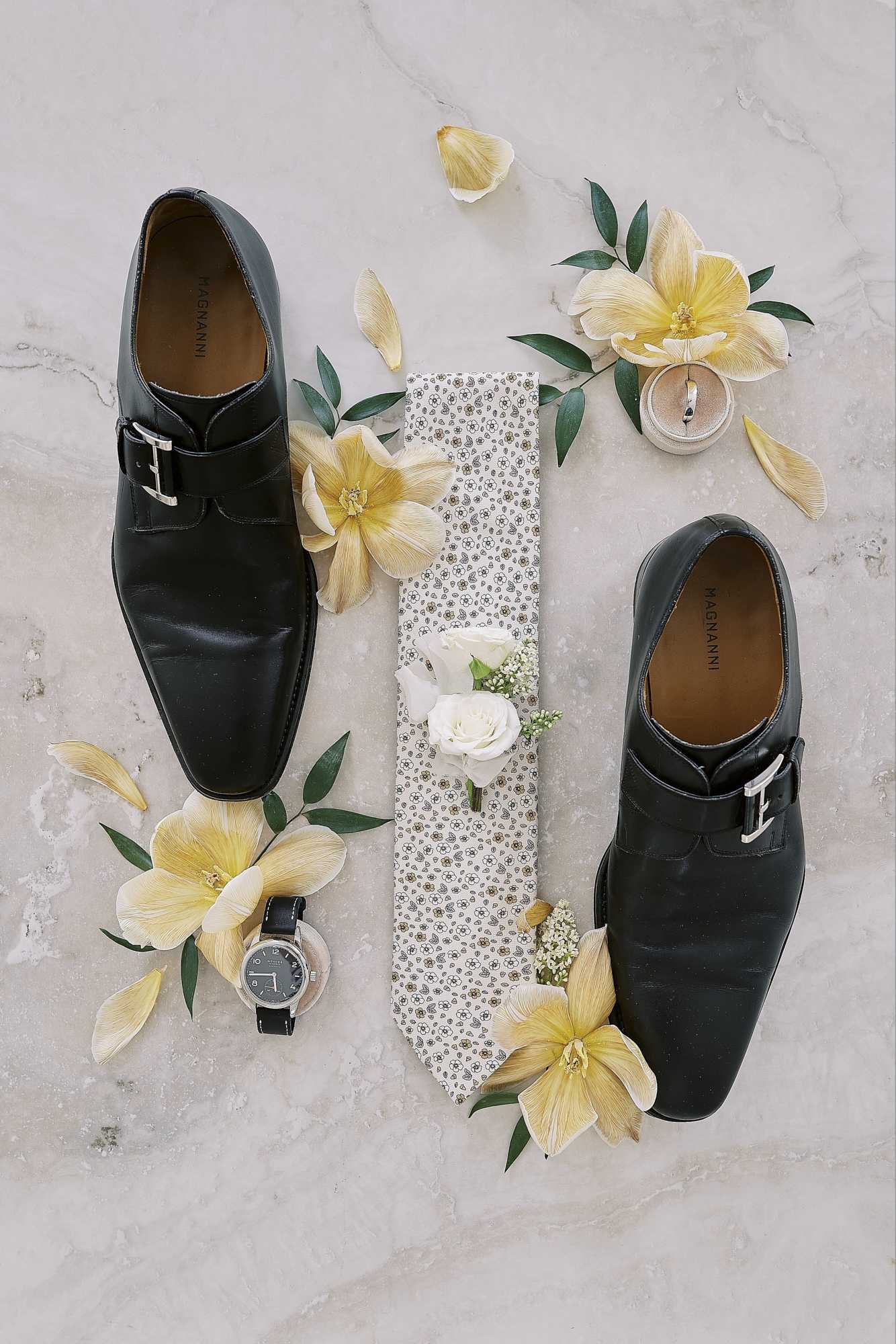 A flat lay detail shot of groom's accessories arranged on a marble surface. The composition includes a pair of black Magnanni single monk-strap dress shoes, a white and grey floral-patterned tie, a white rose and baby's breath boutonniere, a dark-faced leather-strap watch, a ring dish holding a wedding band, and scattered cream and yellow orchid blooms with green foliage as decorative styling elements throughout the arrangement.