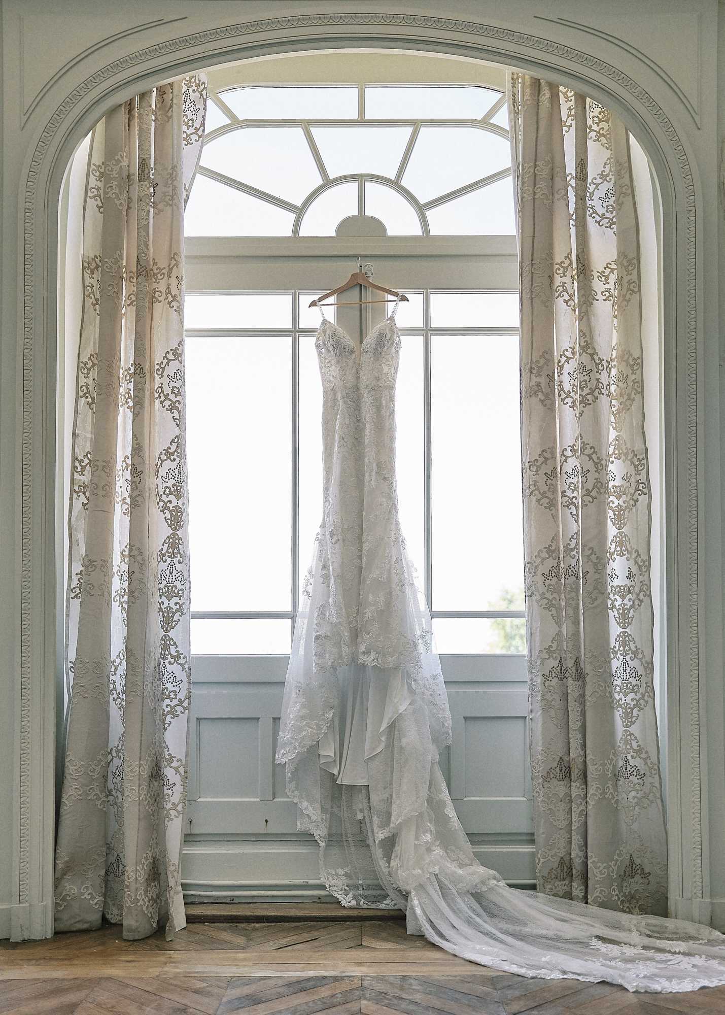 A getting-ready detail shot of an ivory lace wedding gown hanging on a wooden hanger in front of a tall arched window inside a French chateau-style room. The dress features a fitted silhouette with spaghetti straps, a sweetheart neckline with lace appliqué bodice, and a long cathedral lace train that pools on the herringbone parquet floor. The window is framed by a carved plaster arch with sage green painted woodwork and floor-length cream curtains printed with a gold damask pattern. Natural backlight from the window illuminates the dress. Wide portrait composition.