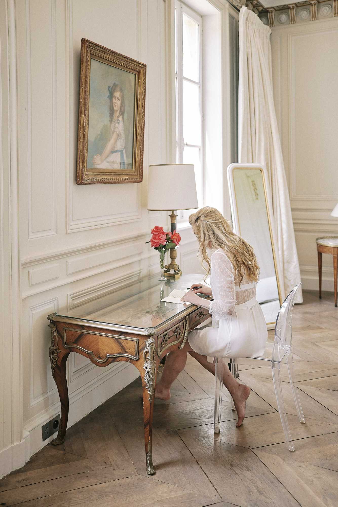 A bride is captured during the getting-ready phase, seated at an ornate Louis XV-style marquetry writing desk with gilded bronze mounts, writing what appears to be wedding vows or a letter. She wears a white lace-sleeved bridal robe or short getting-ready outfit, is barefoot, and has long wavy blonde hair worn down. She sits on a clear acrylic ghost chair. The indoor setting is a classically decorated French chateau room featuring cream boiserie wall paneling, a herringbone parquet floor, ivory floor-length curtains, a gold-framed portrait painting of a girl on the wall, a table lamp, a vase of coral-red roses on the desk, and a gold-framed floor mirror leaning against the wall. The shot is a medium-wide portrait taken from behind and to the side, emphasizing both the bride and the period interior. Potential venue feature image.
