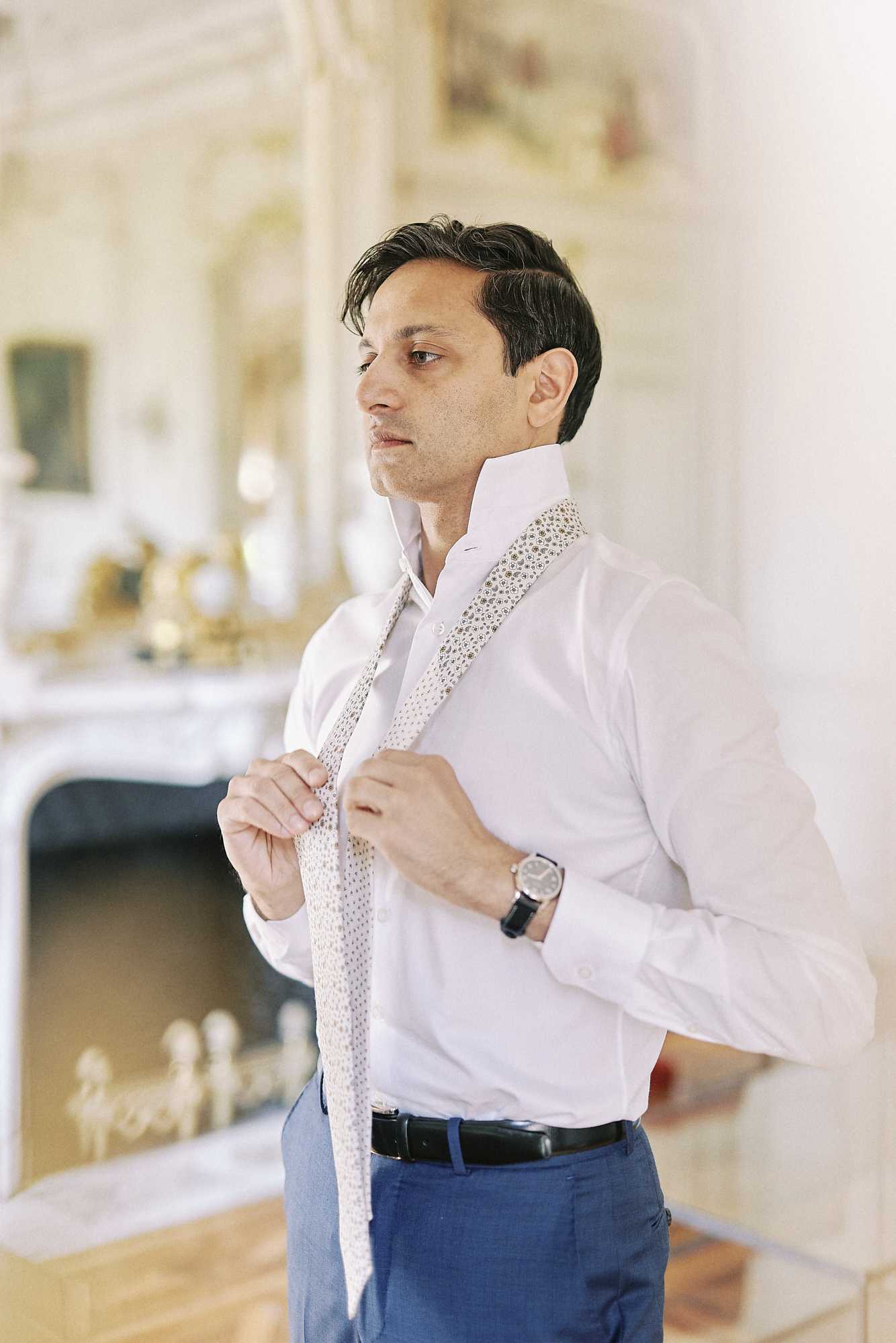 A groom getting ready indoors, adjusting a cream and gold patterned tie around the collar of his white dress shirt. He is wearing cobalt blue trousers with a black leather belt and a classic silver-faced watch with a dark strap. The setting appears to be an ornate chateau room featuring a white marble fireplace with gilded decorative elements, a large mirror with a gold frame, and painted ceiling moldings visible in the background, suggesting a classic French interior. The shot is a medium portrait, slightly off-center, with a soft, airy light tone throughout.