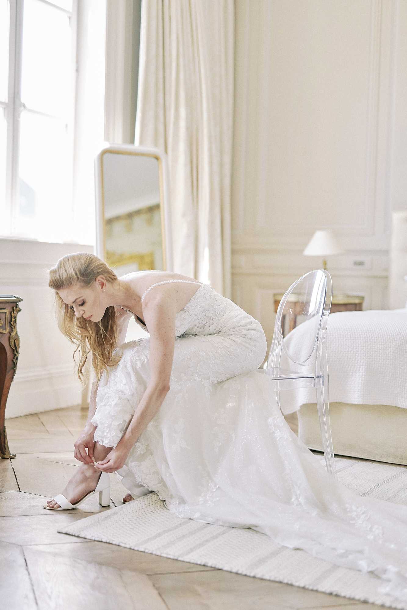 A bride is getting ready indoors, seated on a clear acrylic ghost chair while fastening a white block-heel sandal. She is wearing a fitted, low-back ivory lace gown with floral appliqué detailing and a delicate pearl or button closure along the back neckline, with her blonde hair worn loosely down. The room has the appearance of a classic French chateau-style interior, featuring herringbone parquet flooring, white paneled walls, cream floor-length drapes, a gold-framed standing mirror, an ornate gilded side table, and a white-covered bed visible in the background. The overall styling is clean and classic, with an all-white and ivory palette throughout the dress, shoes, and room furnishings, shot as a full-length portrait with soft natural window light.