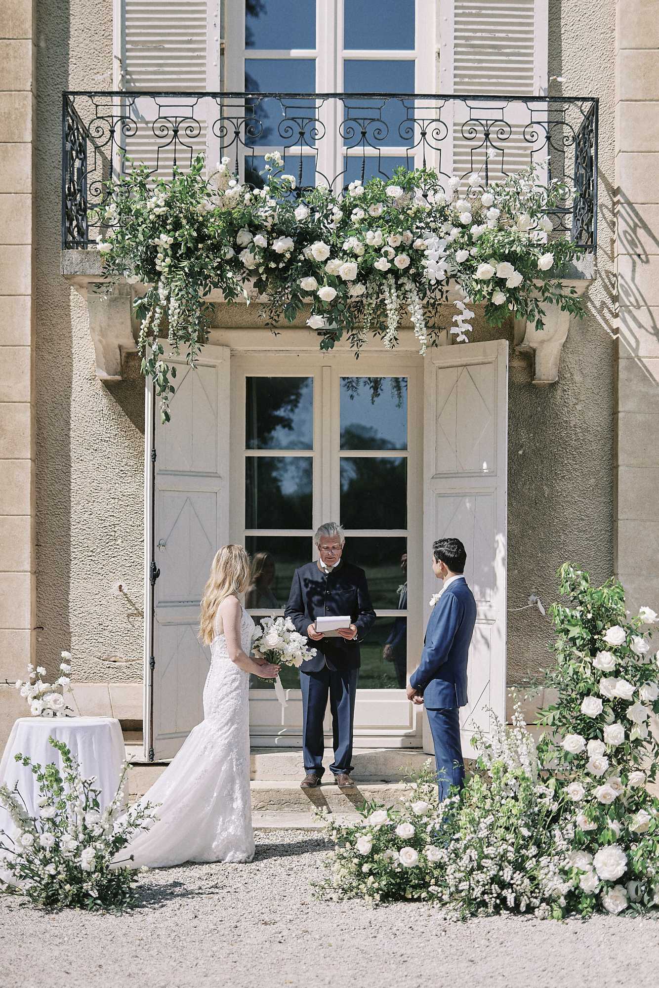 An outdoor wedding ceremony taking place directly in front of the main entrance doors of a French chateau, with the couple standing before an officiant who is reading from a booklet. The bride wears a fitted lace gown with a train and holds a bouquet of white roses and greenery, while the groom is dressed in a navy blue suit. Large ground-level floral installations of white roses, ranunculus, and trailing greenery frame both sides of the doorway, and the wrought-iron balcony above is heavily decorated with cascading white roses, wisteria, and lush greenery. A small white-clothed table is visible to the left. The composition is a wide portrait shot capturing the full ceremony setup against the chateau facade. Potential venue feature image.