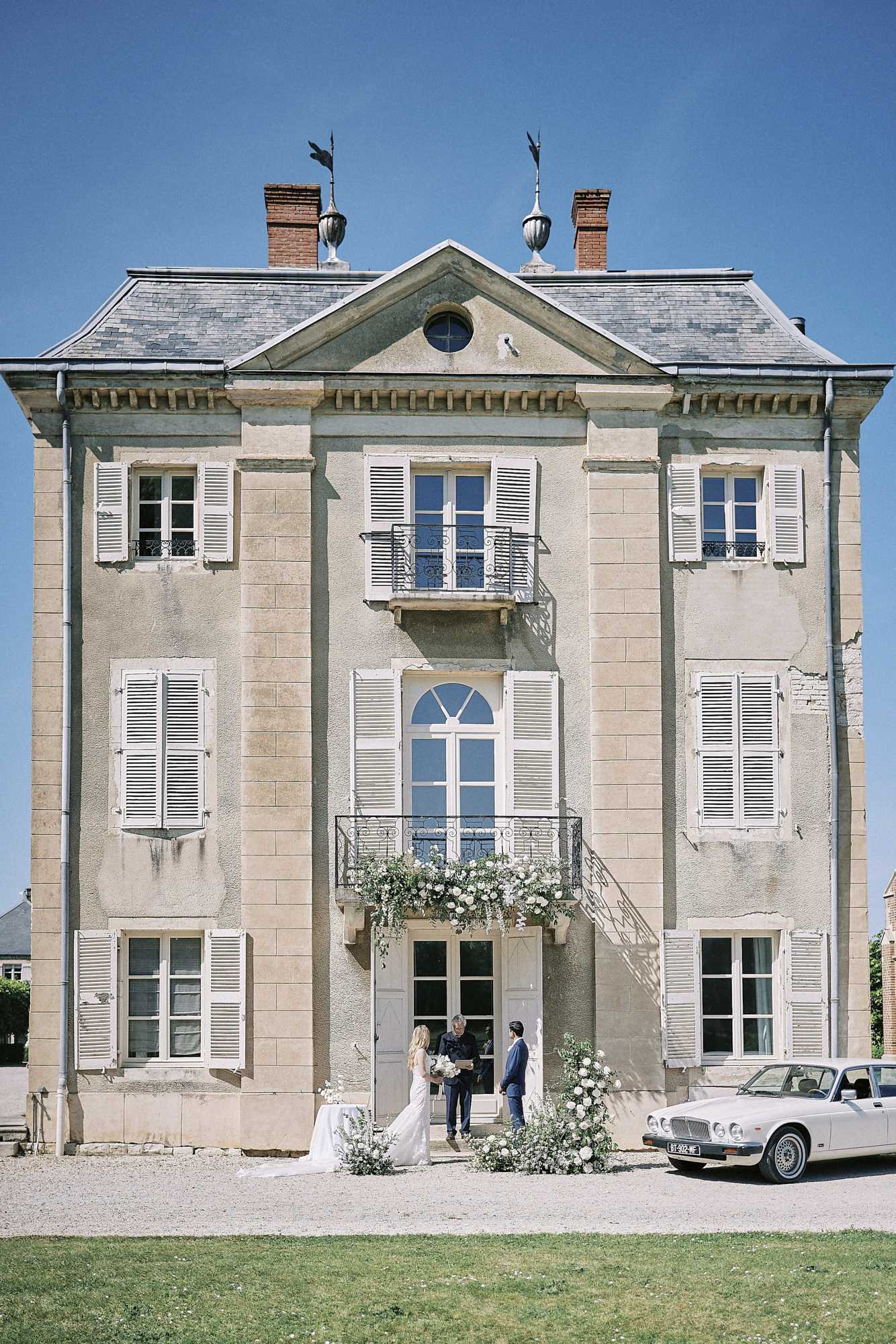 A wide shot of a three-story French château with cream limestone facades, white shuttered windows, a slate mansard roof with decorative finials, and wrought-iron balconies. At the entrance steps, a small wedding ceremony is taking place with three figures visible: a bride in a white fitted gown, a groom in a navy suit, and an officiant in dark attire, all standing beneath a lush floral arch of white blooms and trailing greenery that also decorates the doorway surround. A vintage cream-colored Jaguar sedan is parked to the right of the entrance, and a small table with white linens is positioned to the left. The overall styling is classic and refined with a white and green floral palette. Potential venue feature image.