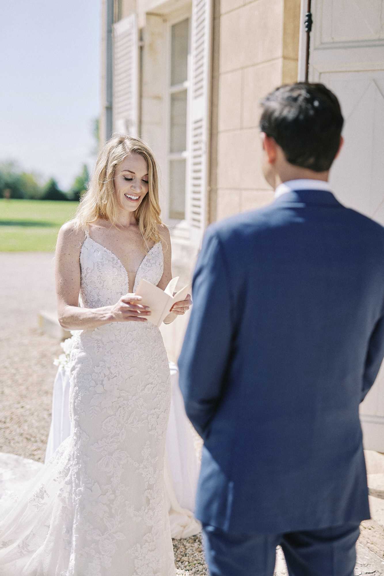 A bride and groom share a private vow exchange outdoors beside the stone facade of a French chateau, likely a first-look or private vow reading moment. The bride, smiling and looking down, reads from a small cream-colored booklet; she wears a fitted ivory lace gown with a deep V-neckline and spaghetti straps, with floral lace detailing throughout the skirt. The groom, seen from behind, wears a navy blue suit. The shot is a medium portrait framing both figures, with the chateau's limestone architecture and white shutters visible in the background.