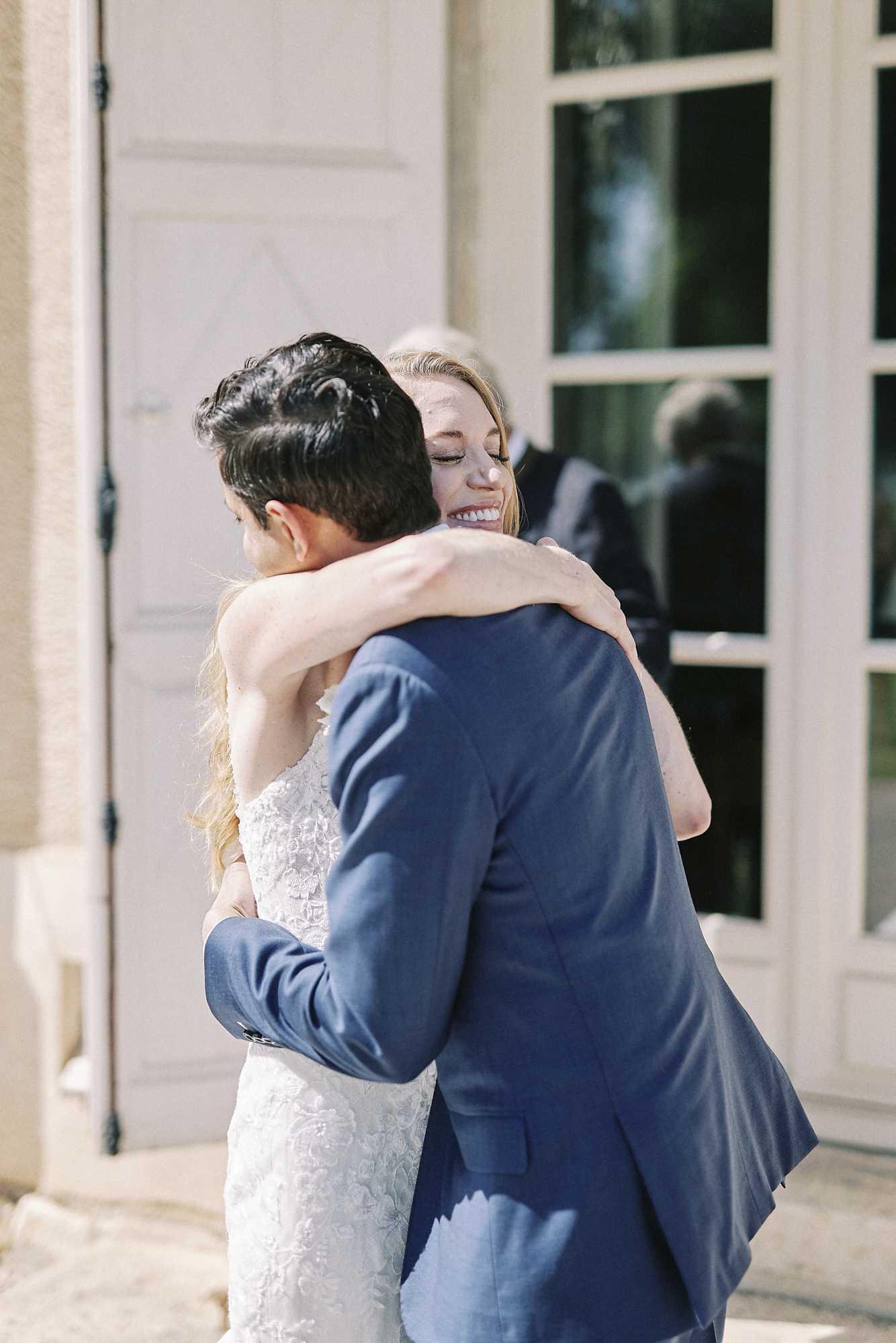 The bride and groom share an embrace outdoors in what appears to be a first look or post-ceremony moment in front of a chateau or manor house entrance. The bride, a blonde woman with long hair, is wearing a strapless fitted lace gown with floral appliqué detail and is smiling broadly with her eyes closed as she wraps her arms around the groom's neck. The groom, seen from behind, is wearing a navy blue suit. A third person, an older individual, is partially visible reflected in the tall French door behind them. The shot is a medium portrait taken from slightly behind the groom, giving focus to the bride's joyful expression.