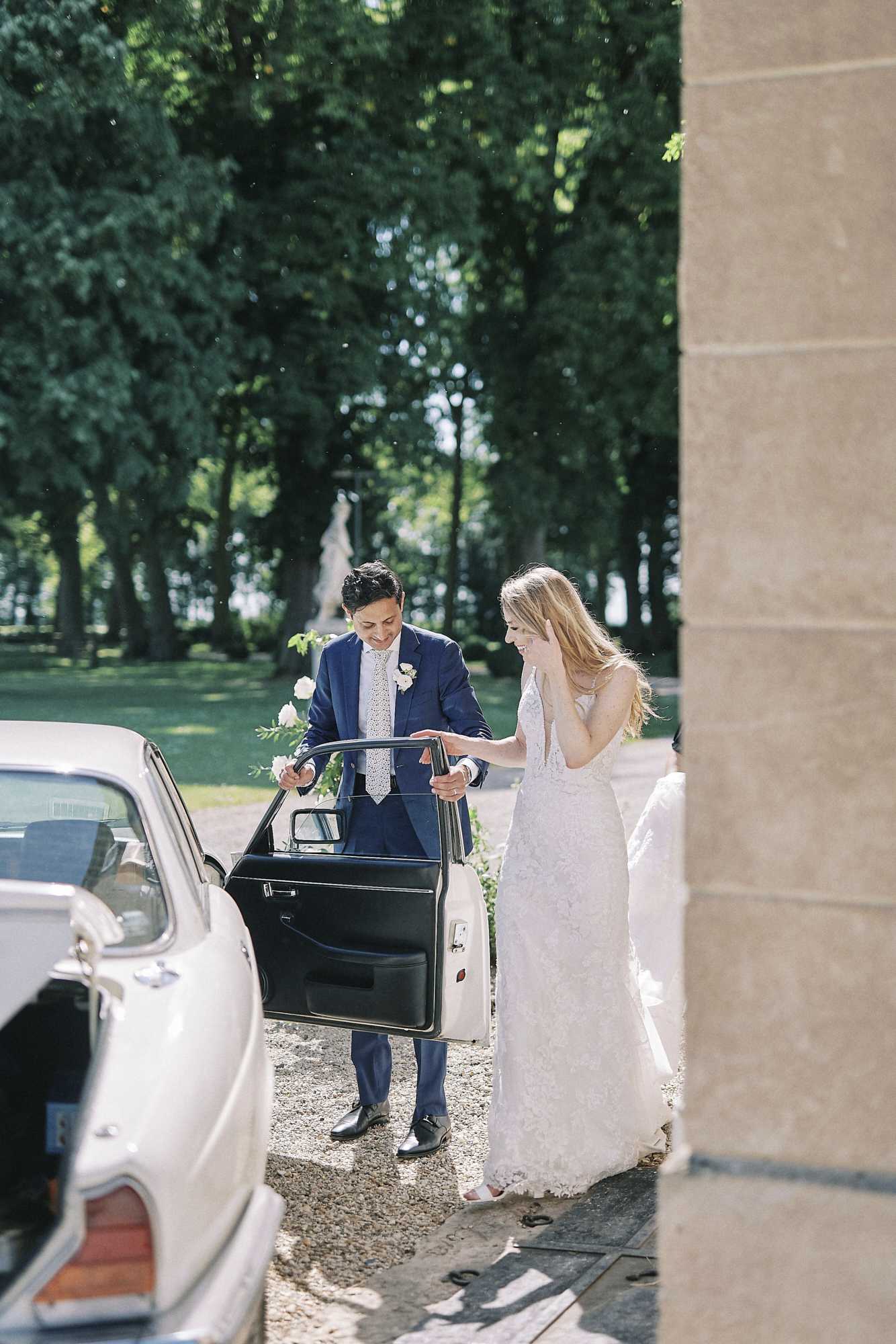 The bride and groom are arriving at or departing from a venue, captured outdoors on a gravel driveway framed by tall trees and a classical stone column. The groom, wearing a navy blue suit with a patterned tie and a white boutonniere, holds the door of a vintage cream-colored car open for the bride. The bride wears a fitted, deep V-neck lace gown with a floral appliqué design and has long blonde hair worn loose; she is smiling and looking down as she steps toward the car. A classical stone statue is visible in the background garden, suggesting a chateau or estate setting. The shot is a medium portrait framed through the column and car door, capturing a candid, relaxed moment between the couple.
