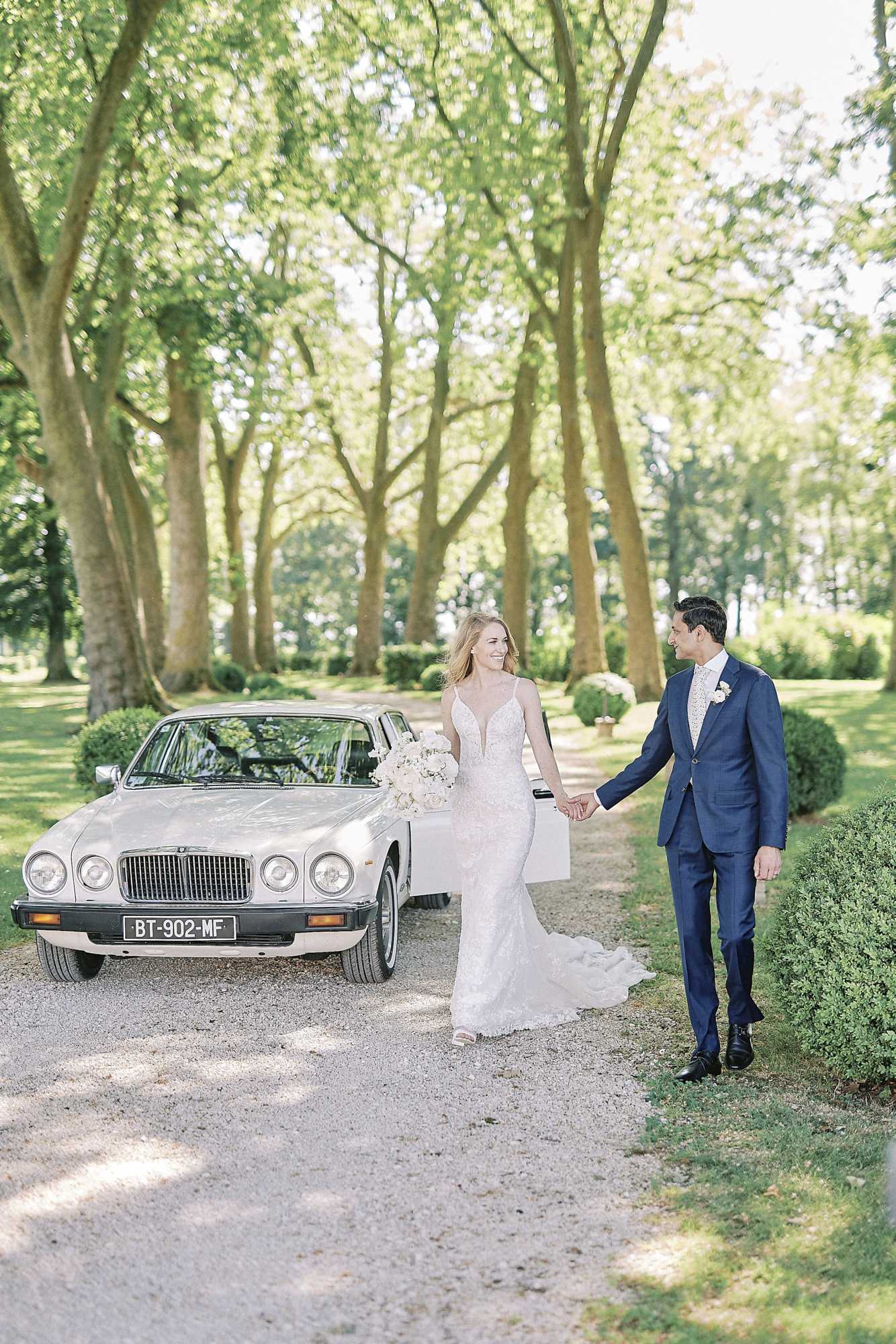 A couple portrait taken outdoors on a gravel tree-lined driveway, with the bride and groom holding hands and walking toward the camera beside a vintage cream Jaguar sedan with a French license plate. The bride wears a fitted lace gown with a deep V-neckline and a short train, carrying a large bouquet of ivory and white blooms including what appear to be peonies and roses. The groom wears a navy blue suit with a floral tie and a white boutonniere. The setting features a formal garden with neatly trimmed boxwood hedges lining the driveway, consistent with a French chateau property. The shot is a full-length portrait with natural daytime lighting filtering through the tree canopy.
