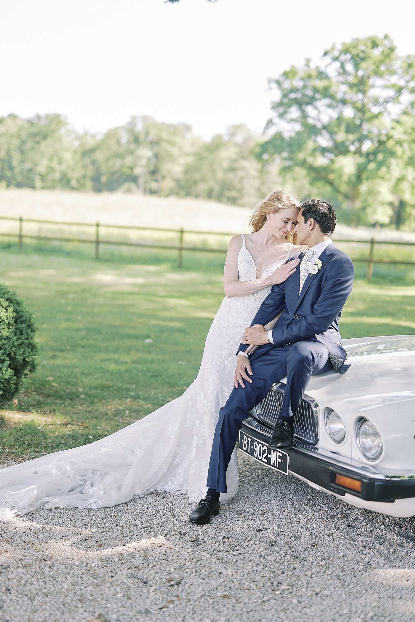 Outdoor couples portrait of a bride and groom posed against the front hood of a vintage white car with a French license plate, set on a gravel driveway with a fenced estate grounds in the background. The groom, wearing a navy blue suit with a patterned bow tie and a white boutonniere, sits on the car's hood while the bride leans in close with their foreheads nearly touching. The bride wears a fitted lace gown with thin spaghetti straps, a deep V-neckline, and a long cathedral-length train that fans out across the gravel. The composition is a full-length portrait shot with soft, bright natural light giving the image an airy, slightly overexposed feel.