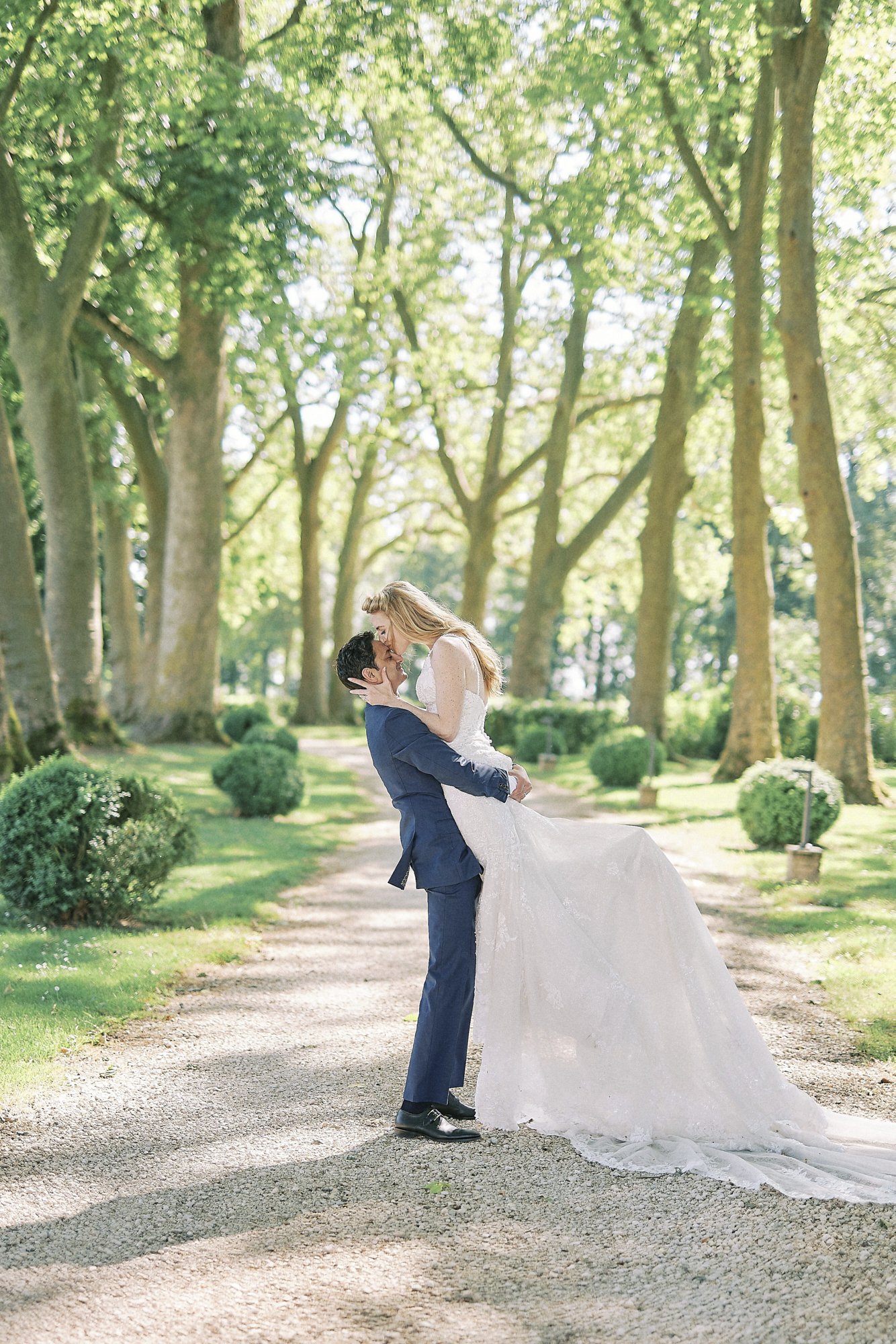 A couple portrait taken outdoors on a tree-lined gravel driveway, where the groom is lifting the bride as they kiss. The groom wears a navy blue suit with black dress shoes, and the bride wears a white lace ballgown with a long cathedral-length train and a halter or spaghetti-strap neckline, her blonde hair worn loose. The avenue is flanked by tall plane trees with pale trunks and a symmetrical row of clipped spherical boxwood shrubs along each side, suggesting a formal French château or domaine garden setting. The shot is a full-length portrait framed centrally within the tree-lined perspective, taken in bright daytime light.