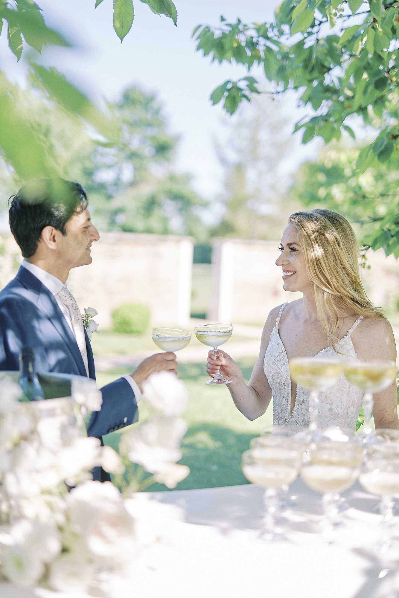 The bride and groom share a champagne toast outdoors in a formal garden setting, clinking coupe-style glasses filled with a light sparkling wine. The groom wears a navy blue suit with a floral tie and a white boutonniere, while the bride wears a white lace spaghetti-strap gown with a deep V-neckline and her blonde hair worn loose. In the foreground, several additional coupe glasses and white florals on a table are softly out of focus, suggesting a cocktail hour setup. The image is a medium portrait shot with a bright, airy quality and a walled garden visible in the background.