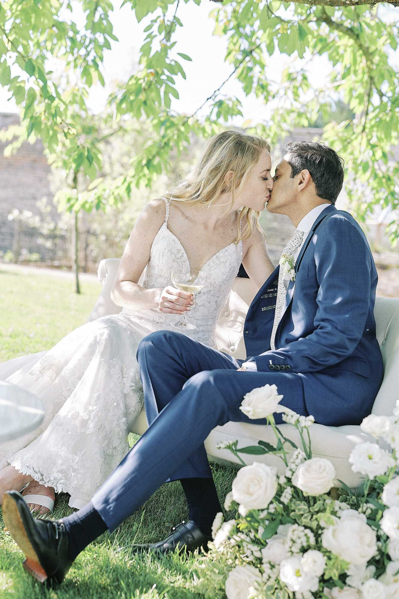 A couple portrait taken outdoors in a garden setting, with the bride and groom seated on a white upholstered loveseat and sharing a kiss. The bride wears a white lace spaghetti-strap gown with a deep V-neckline and holds a coupe glass of champagne, while the groom wears a navy blue suit with a patterned tie and a small white floral boutonniere. A large arrangement of white ranunculus, roses, and small white filler flowers is positioned at the base of the loveseat in the foreground. The image is a medium portrait shot with soft natural light filtering through tree branches overhead.