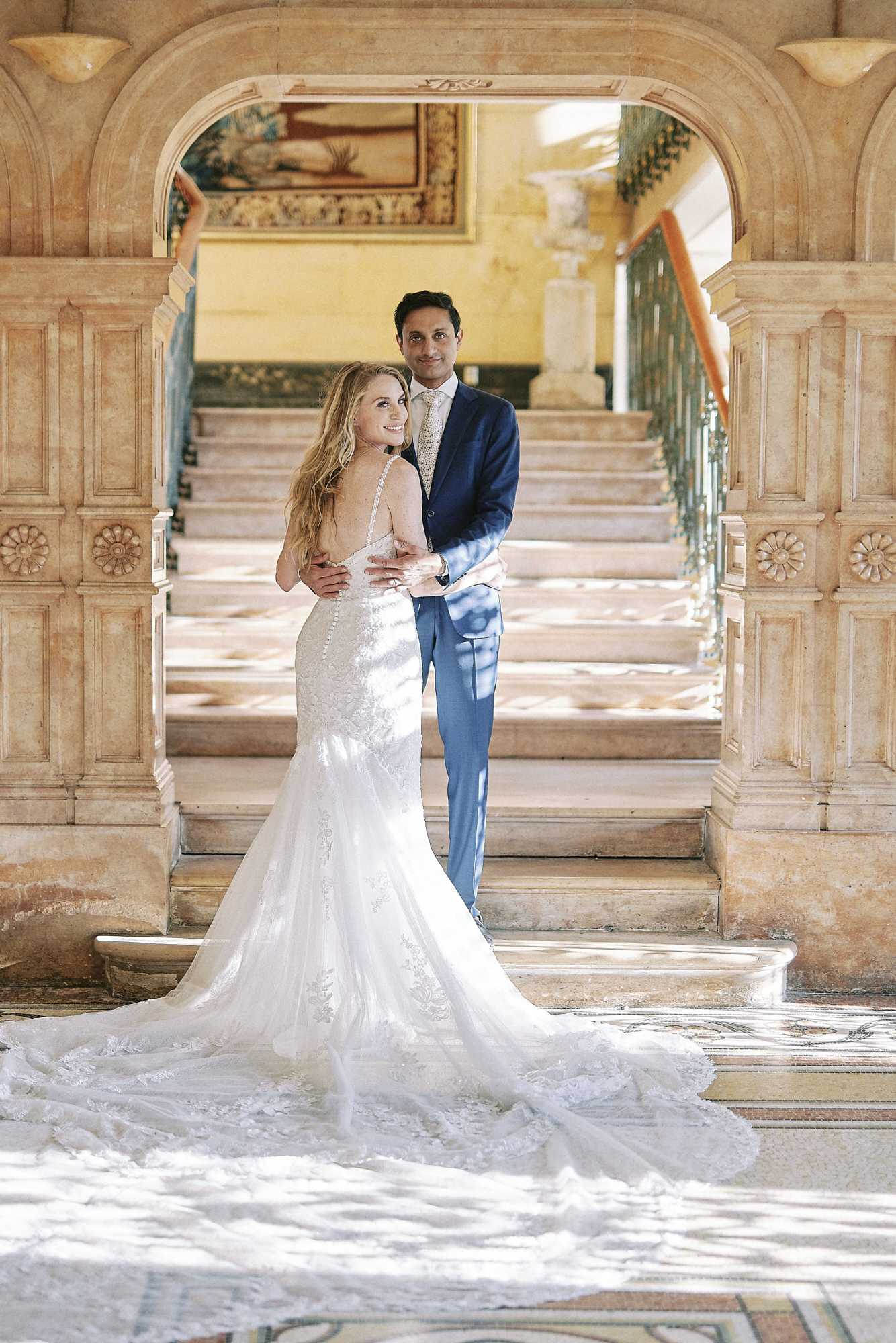 A couple portrait taken indoors at what appears to be a chateau or grand manor, positioned at the base of an ornate marble staircase framed by a carved stone archway with decorative pilasters and floral relief details. The bride wears a fitted ivory lace mermaid-style gown with spaghetti straps, a low open back with button detailing, and a long lace-appliqué train that fans across the tiled floor; she is turned away from camera and looks back over her shoulder. The groom stands behind her with his hands at her waist, wearing a navy blue suit with a patterned gold tie. A large framed painting is visible on the wall at the top of the staircase, and the interior features ornate ironwork balusters and warm stone tones throughout. Full-length couple portrait shot.