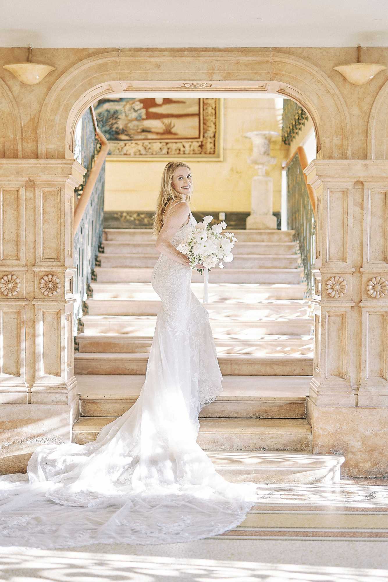 A bridal portrait taken indoors at the base of a grand marble staircase framed by an ornate carved stone archway with decorative rosette panels and wall sconces. The bride stands on the bottom step, turned slightly away from camera and glancing back with a smile, wearing a fitted lace mermaid-style gown with spaghetti straps and an extended lace train that spreads across the floor. She holds a rounded bouquet of white ranunculus and garden roses with a white ribbon. A large framed tapestry painting and wrought-iron balustrade are visible in the background through the arch. The image is a full-length portrait shot in bright, airy natural light. Potential venue feature image.