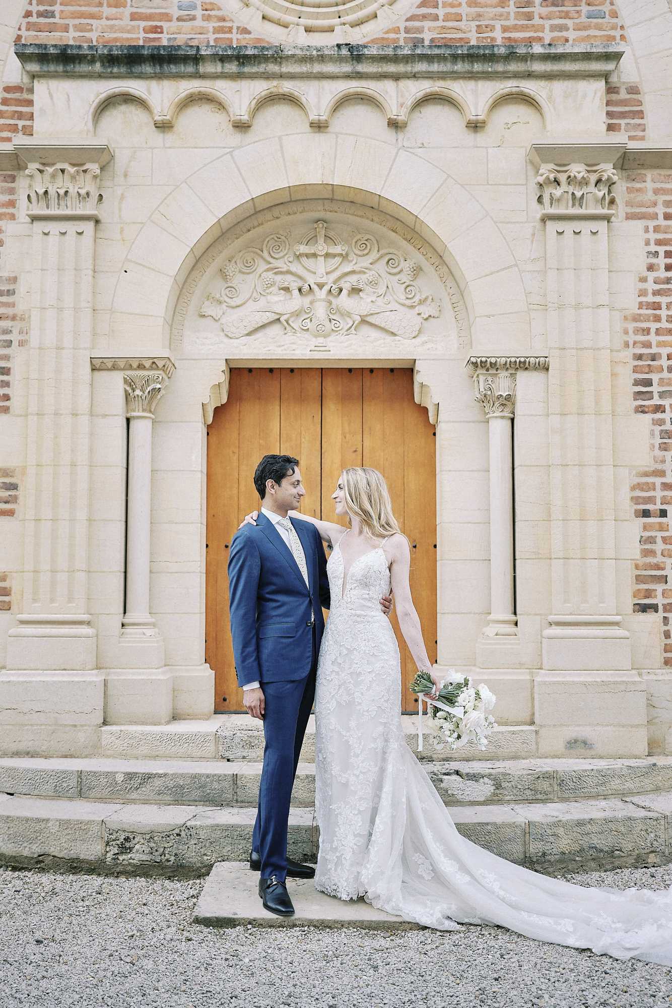 A couple portrait taken outdoors in front of an ornate chapel or church entrance featuring a carved stone archway with decorative relief work, flanked by stone columns, and large wooden double doors. The groom wears a navy blue suit with a light grey tie and black dress shoes, while the bride wears a fitted lace gown with spaghetti straps, a deep V-neckline, and a long train. She holds a bouquet of white flowers with greenery and has her arm around the groom's shoulder as they look at each other. The composition is a full-length couple portrait with the architectural facade as a prominent backdrop. Potential venue feature image.