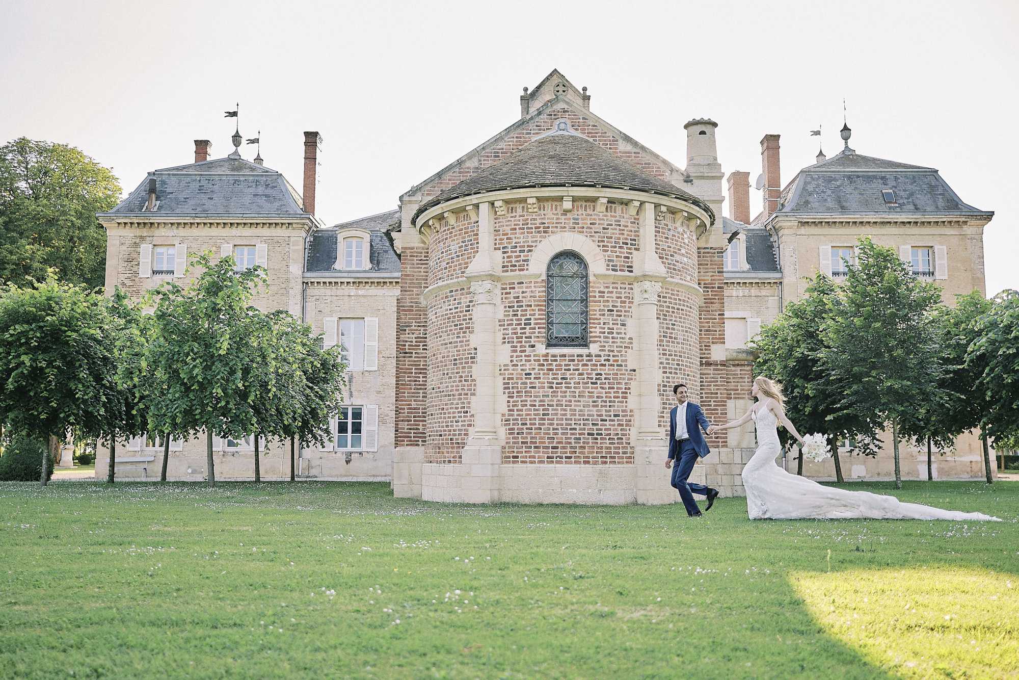 A couple portrait taken outdoors on the grounds of a French chateau, with the bride and groom walking and holding hands across a wide lawn in front of the main building. The groom wears a navy blue suit, and the bride wears a fitted ivory lace gown with a long cathedral-length train; she carries a white bouquet, likely large-headed blooms such as peonies or hydrangeas. The chateau features a distinctive rounded brick apse at its center with a leaded arched window, flanked by two symmetrical stone wings with slate mansard roofs and red brick chimneys. The composition is a wide shot that places the couple small within the frame, emphasizing the scale and architecture of the venue. Potential venue feature image.