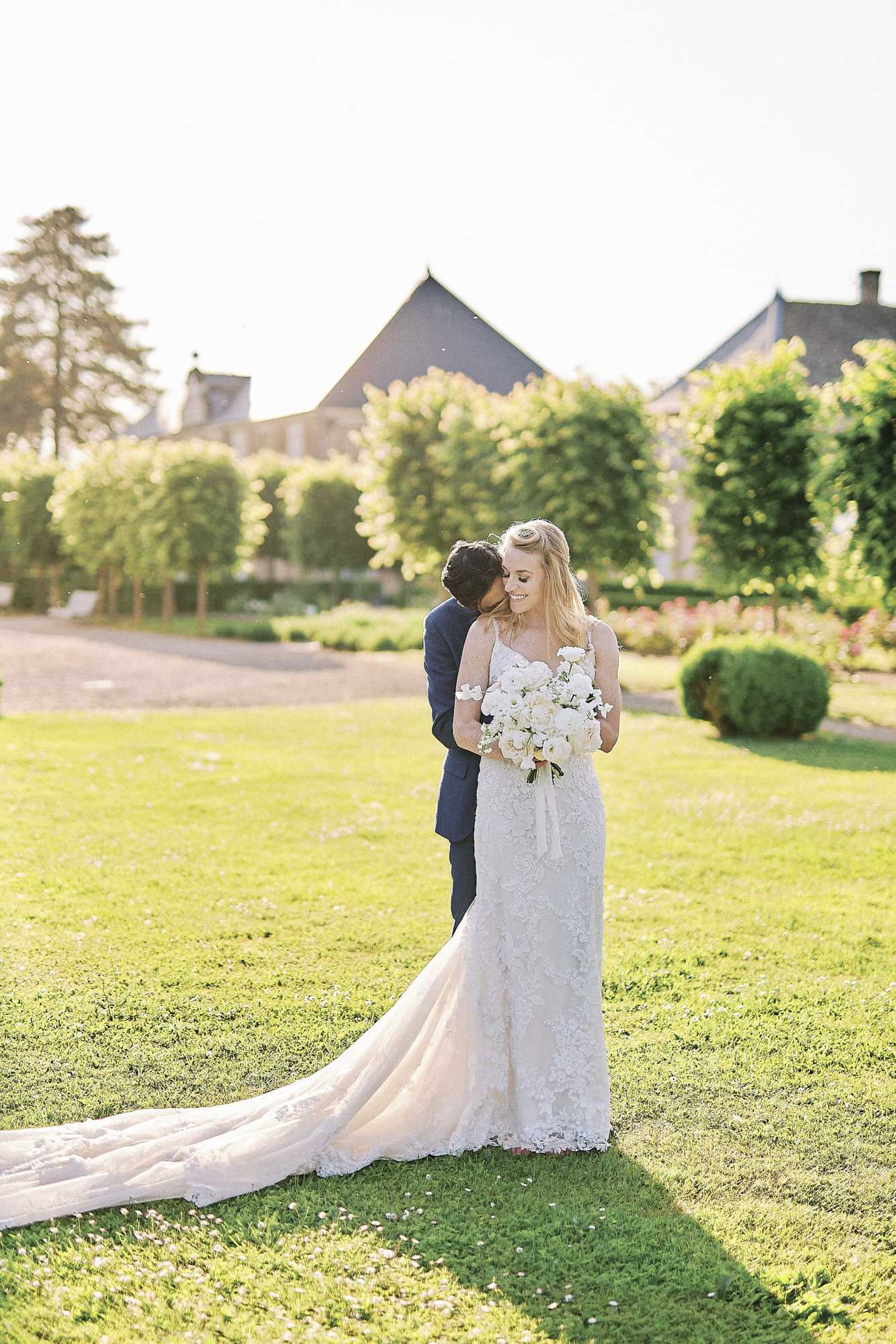 A couple portrait taken outdoors in the formal garden of what appears to be a French chateau or abbey, with stone buildings visible in the soft-focus background. The bride wears a fitted ivory lace gown with thin spaghetti straps and a long cathedral train spread across the lawn, and holds a full bouquet of white ranunculus and garden roses. The groom, dressed in a navy suit, stands behind the bride nuzzling her cheek while she smiles downward. The shot is a medium full-length portrait taken in warm golden-hour light, with manicured hedges and topiary visible in the background.