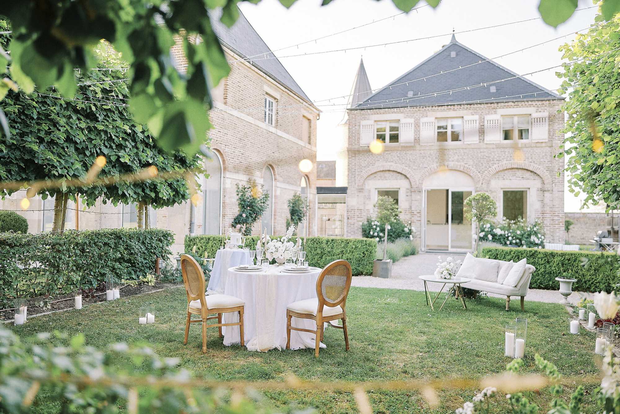An outdoor reception setup photographed from a medium-wide angle through foreground foliage at dusk, set in the manicured garden courtyard of a classic French brick manor house. A sweetheart table for two is centered on the lawn, dressed in a white linen tablecloth with gold cane-back Louis XVI-style chairs, topped with a white floral centerpiece of roses and orchids, crystal glassware, and taper candles. Pillar candles in glass hurricane holders are scattered across the lawn, and a cream upholstered loveseat with a gold-frame coffee table is positioned to the right as a lounge area. Fairy lights are strung overhead across the courtyard, and the formal hedgerow parterre garden and pale stone manor facade with arched windows serve as the backdrop. The overall decor palette is white and gold with a classic, refined French aesthetic. Potential venue feature image.
