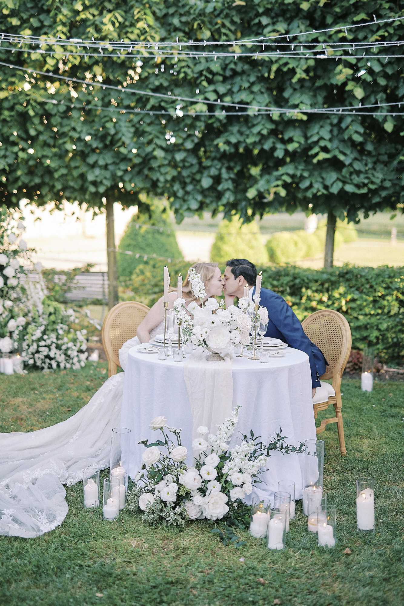 A bride and groom share a kiss at an outdoor sweetheart table set within a formal garden, with fairy lights strung overhead and manicured hedges and trees in the background. The round table is dressed with a white linen cloth and a white table runner, topped with a dense centerpiece of white ranunculus, roses, and stock flowers in green foliage, flanked by tall taper candles in gold candlesticks. The bride wears a white gown with a long lace-edged train that pools on the lawn, and the groom is dressed in a navy blue suit; they are seated in gold cane-back chairs. The base of the table and surrounding lawn are decorated with a ground-level floral arrangement of white roses, ranunculus, and greenery, surrounded by numerous white pillar candles in glass hurricane holders, creating a classic, all-white decor palette. Wide portrait shot.