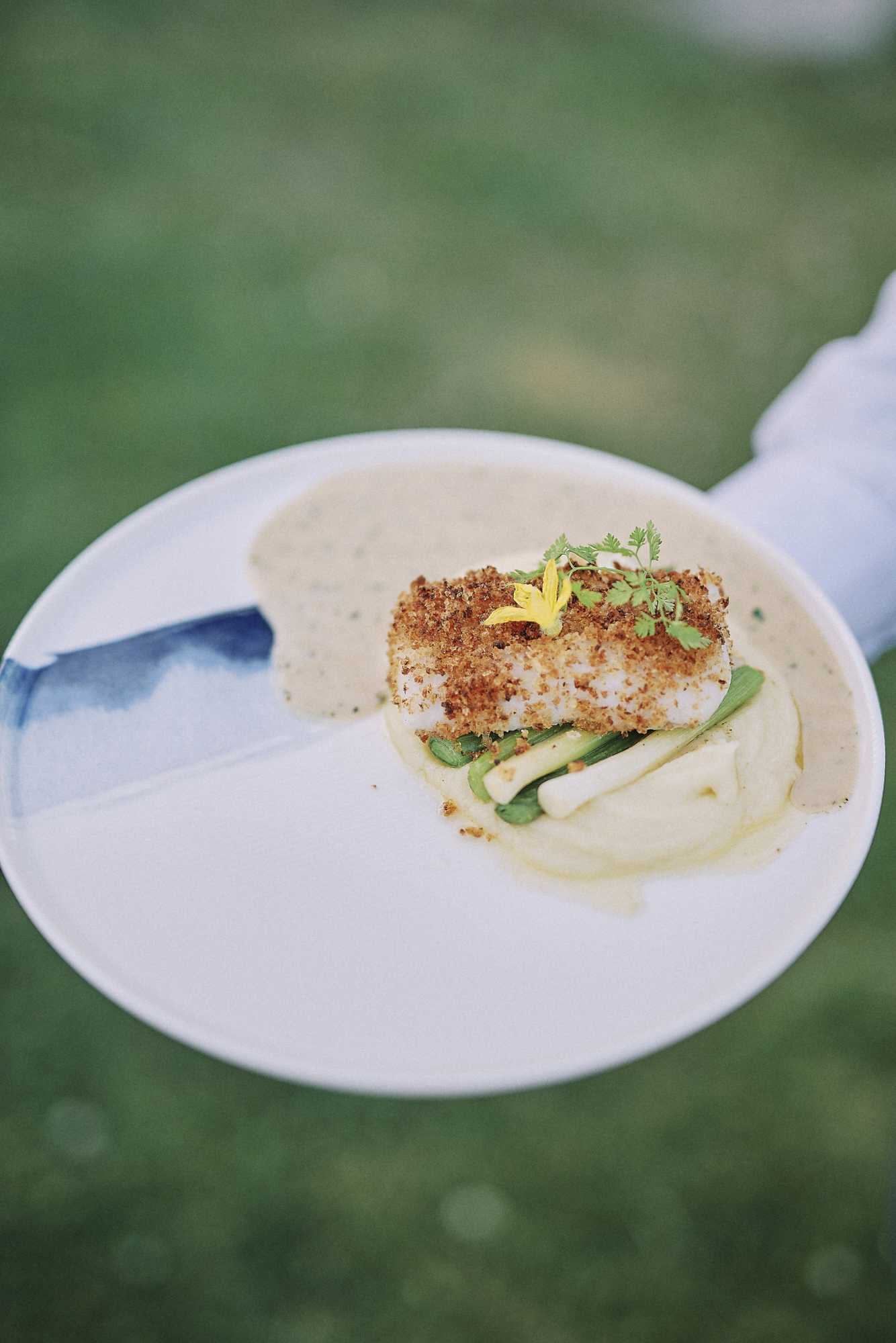 A close-up detail shot of a plated wedding reception dinner course being held by a server in a white uniform against an outdoor green background. The white ceramic plate features a blue watercolor-style brushstroke detail and holds a breadcrumb-crusted fish fillet served over a pale purée, accompanied by green beans and white asparagus spears, garnished with a small yellow edible flower and fresh herb micro-greens. The composition is a tight overhead-angled close-up with a shallow depth of field, emphasizing the modern, refined plating style of the catering.