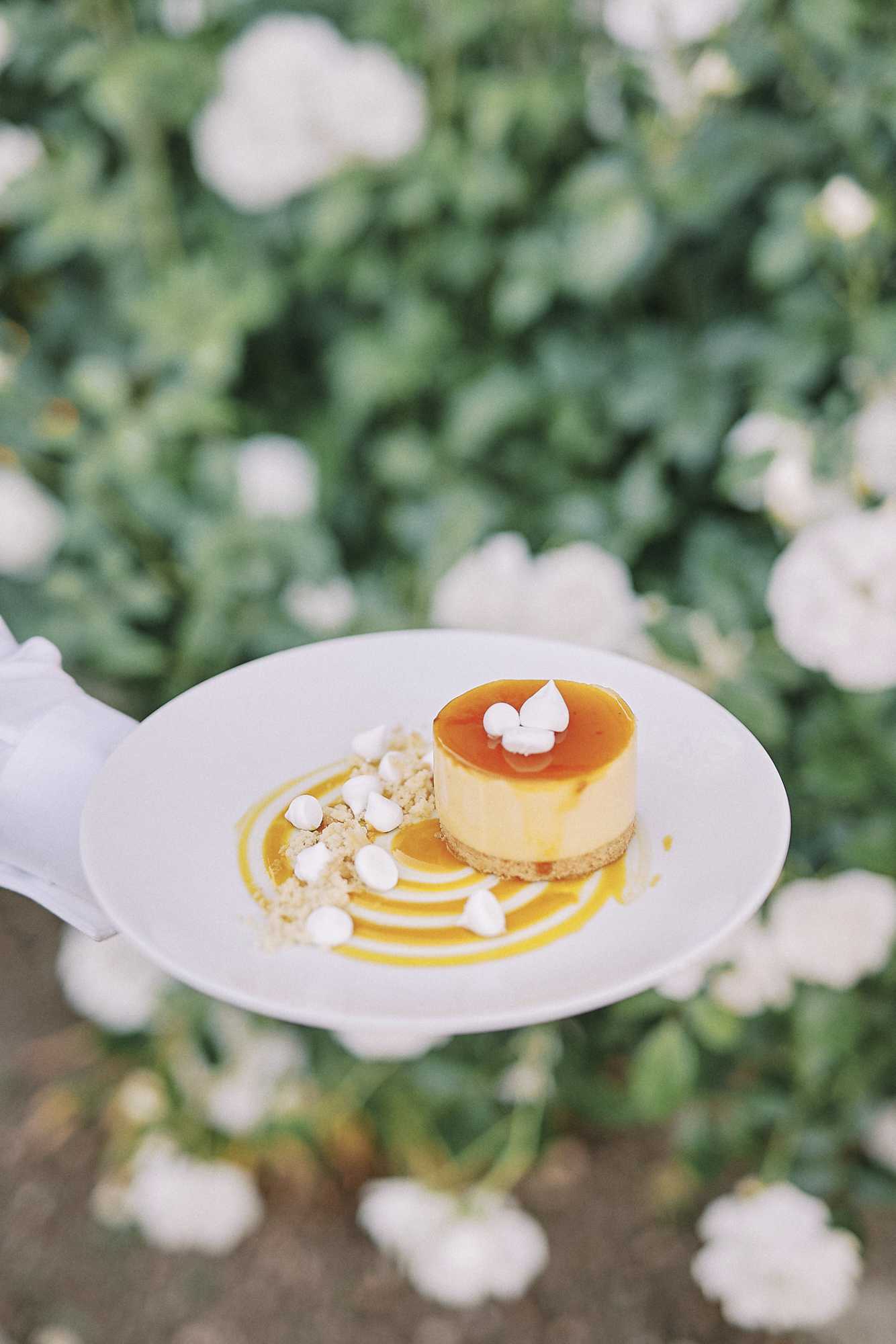 A close-up detail shot of a plated individual dessert being held by a white-gloved server's hand against an outdoor backdrop of blurred white blooms. The dessert is a small round individual cheesecake or mousse with a biscuit base, topped with an amber caramel glaze and three small white meringue peaks. The white plate is decorated with a swirl of golden-yellow sauce, crumbled biscuit, and small white meringue drops. The presentation style is modern and refined, consistent with a formal wedding reception catering service.