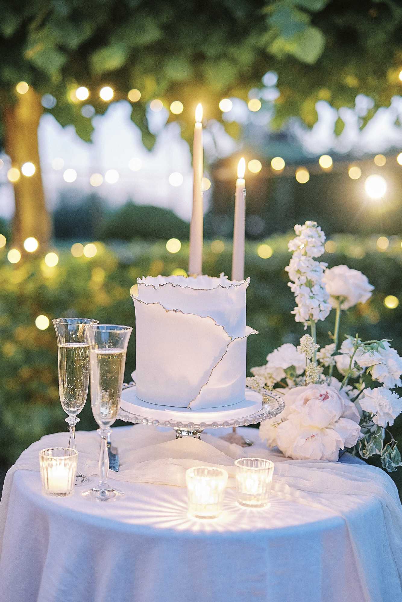A close-up detail shot of an outdoor wedding cake table at dusk, set beneath trees strung with warm globe lights. The table is covered with a white linen and holds a single-tier white wedding cake with a torn-edge fondant design and gold trim detailing, displayed on a silver beaded cake stand with two lit tapered ivory candles inserted into the top. Two champagne flutes filled with sparkling wine are placed to the left of the cake, and three lit glass votive candles are arranged at the front of the table. A loose floral arrangement of blush peonies, white stock, and small white blooms is positioned to the right of the cake. The overall decor palette is white, blush, and gold with a classic, romantic styling approach.