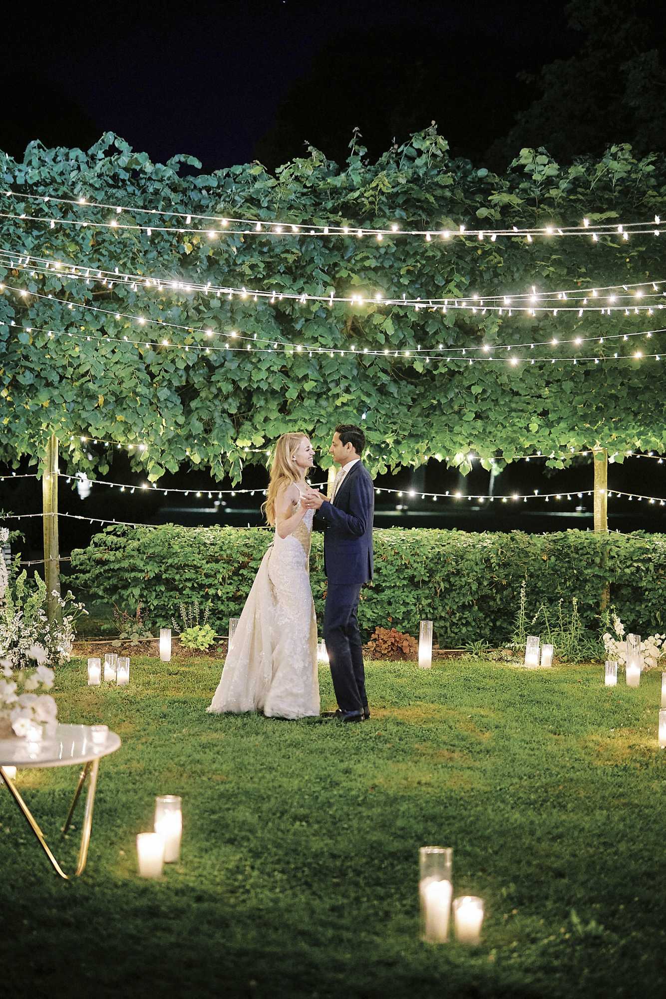 A couple shares their first dance outdoors at night on a garden lawn, surrounded by dozens of lit pillar candles in glass hurricane holders arranged across the grass. The bride wears a fitted ivory lace gown with a flowing skirt, and the groom is dressed in a navy suit. Multiple strands of bistro fairy lights are strung overhead between wooden posts, illuminating a backdrop of dense green climbing vines and a trimmed hedge. A small gold-legged side table with white florals is visible at the left edge of the frame. The wide shot captures the full scene with warm candlelight glowing against the dark night sky, giving the outdoor space a romantic, garden-party atmosphere.