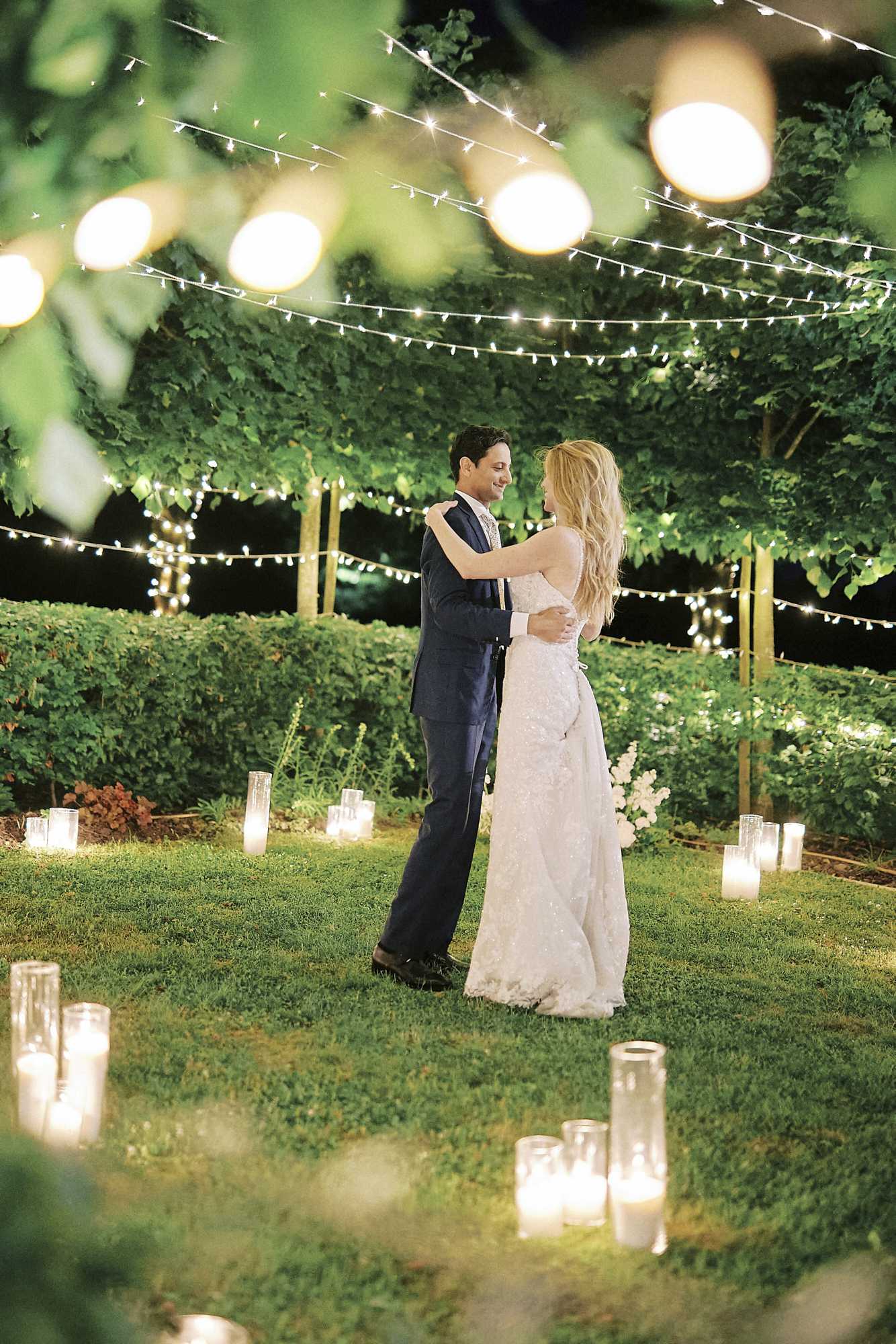 A couple shares a first dance outdoors at night in a garden setting, surrounded by tall glass hurricane candle holders placed directly on the lawn in a loose circular arrangement. The bride wears a fitted white lace gown with a short train, and the groom wears a navy suit with a patterned tie. Overhead, multiple strands of fairy lights and larger globe string lights are strung between trees, creating warm overhead illumination. The garden backdrop features manicured hedges and trellised trees lit from below. The shot is a full-length wide portrait taken from a slight distance, framed by out-of-focus foliage in the foreground, giving depth to the composition.