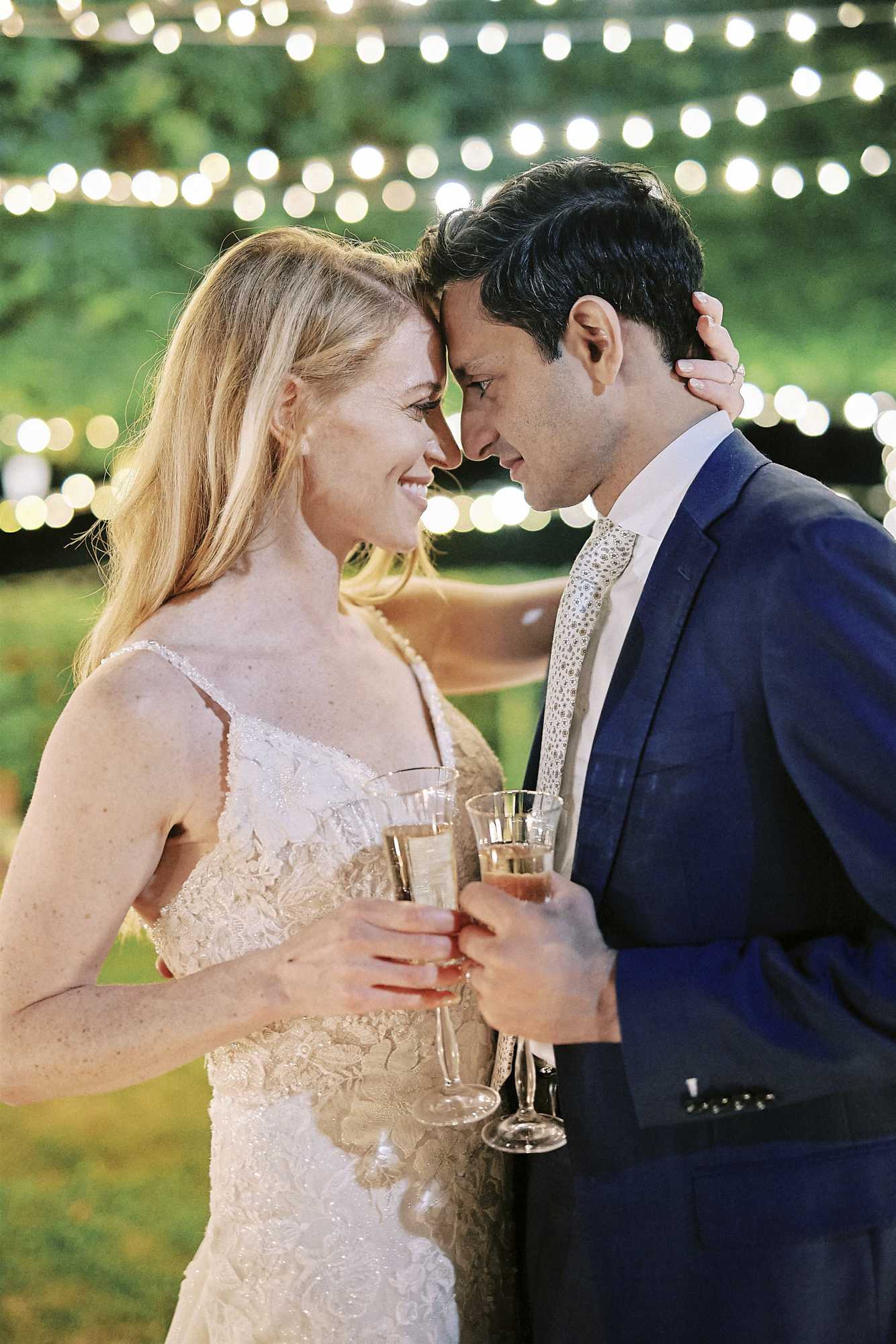 A close-up portrait of a bride and groom outdoors at an evening reception, their foreheads touching as they smile at each other. The bride wears a spaghetti-strap lace gown with floral appliqué detailing, and the groom is dressed in a navy blue suit with a patterned champagne-toned tie. Both are holding champagne flutes, and the bride's hand rests on the back of the groom's neck. The background features strands of warm globe fairy lights draped overhead, creating a soft, glowing bokeh effect against greenery.