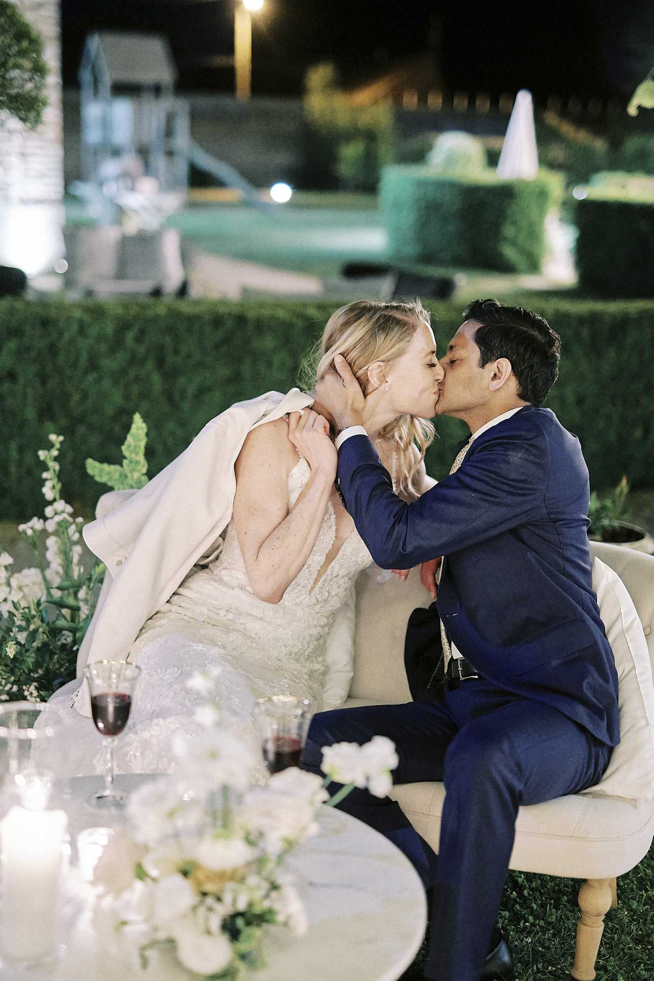 The bride and groom share a kiss while seated together on a cream upholstered loveseat during an outdoor evening reception, likely in the garden of a French chateau or estate. The bride wears a fitted lace gown with a blush-toned jacket draped over her shoulders, while the groom wears a navy blue suit with a gold tie. In the foreground, a small round table holds two glasses of red wine, lit candles, and a low arrangement of white flowers including what appears to be white ranunculus and green amaranthus stems. The background shows manicured box hedges and exterior lighting, giving a classic formal garden setting at night. Portrait-style shot with soft foreground blur.